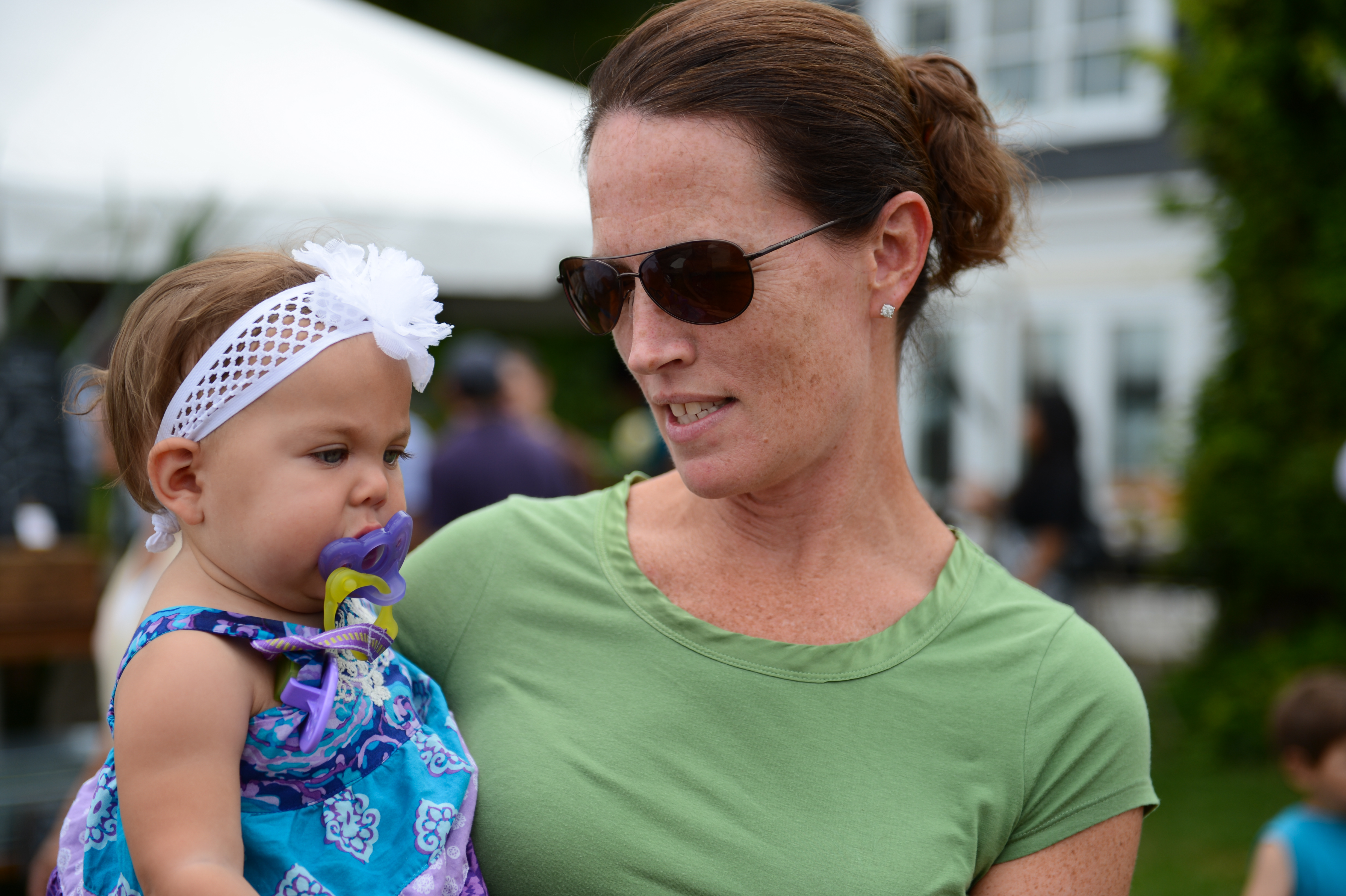 A woman in sunglasses holds a baby with a pacifier at an outdoor gathering.
