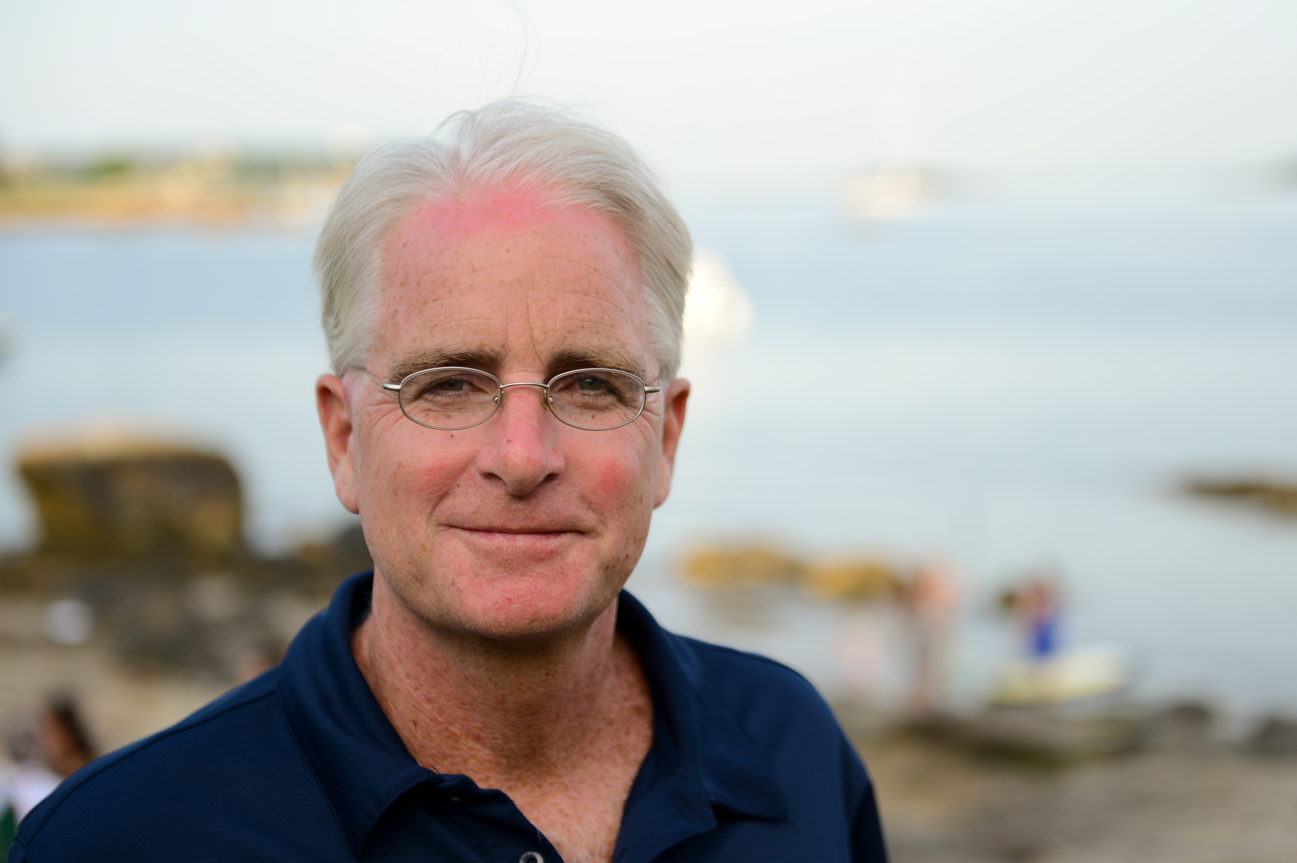 A man in glasses and a navy shirt smiles at the camera with a beach setting in the background.