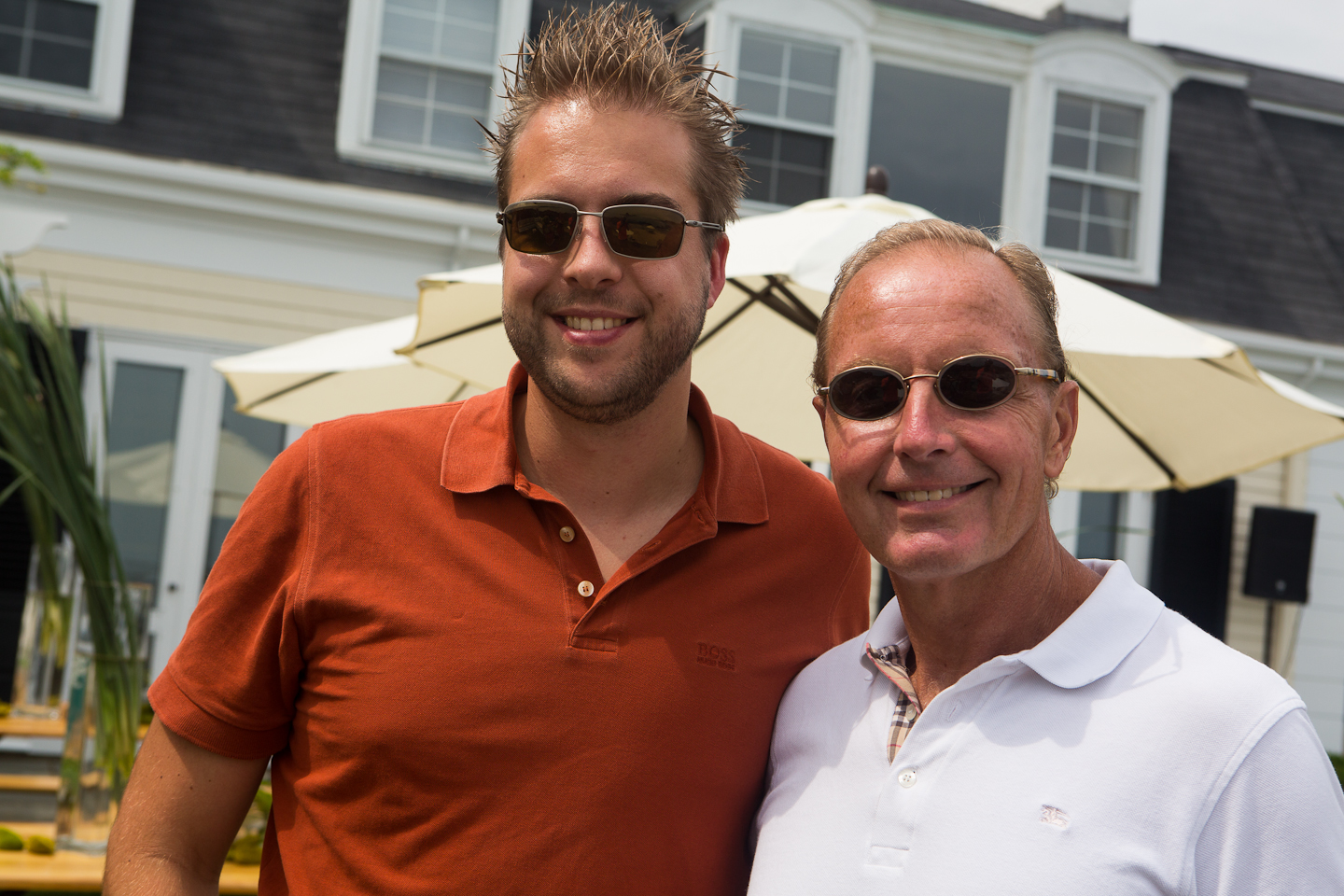 Two men wearing sunglasses smile at the camera while standing outdoors at a summer party.