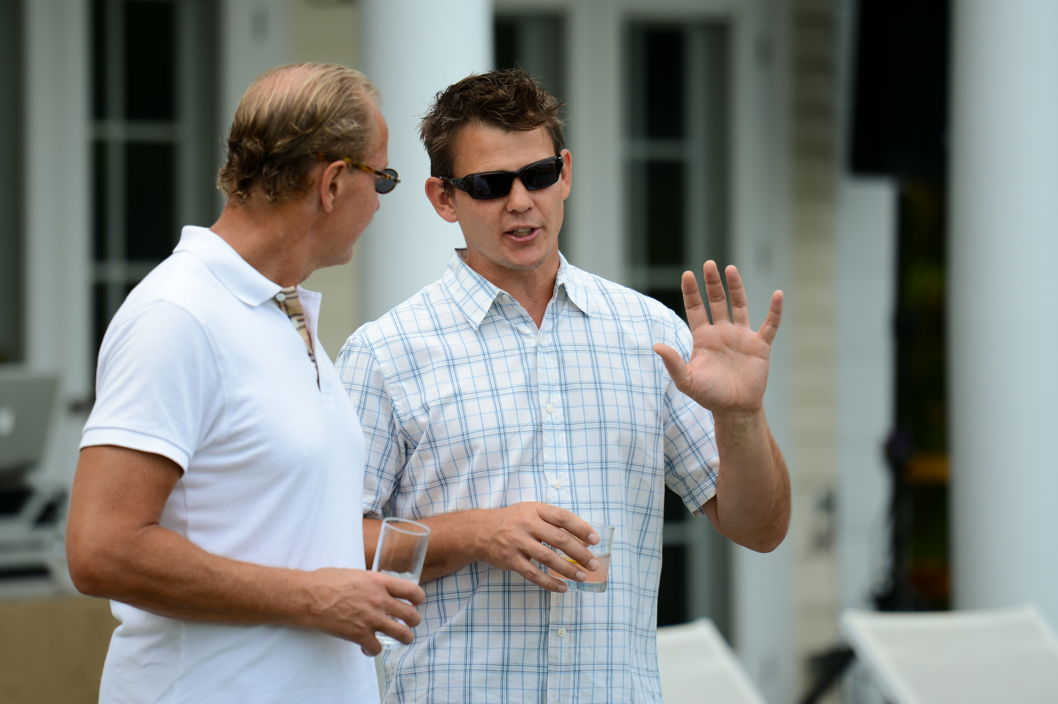 Two men wearing sunglasses talk outside, one gesturing with his hand while the other holds a drink.