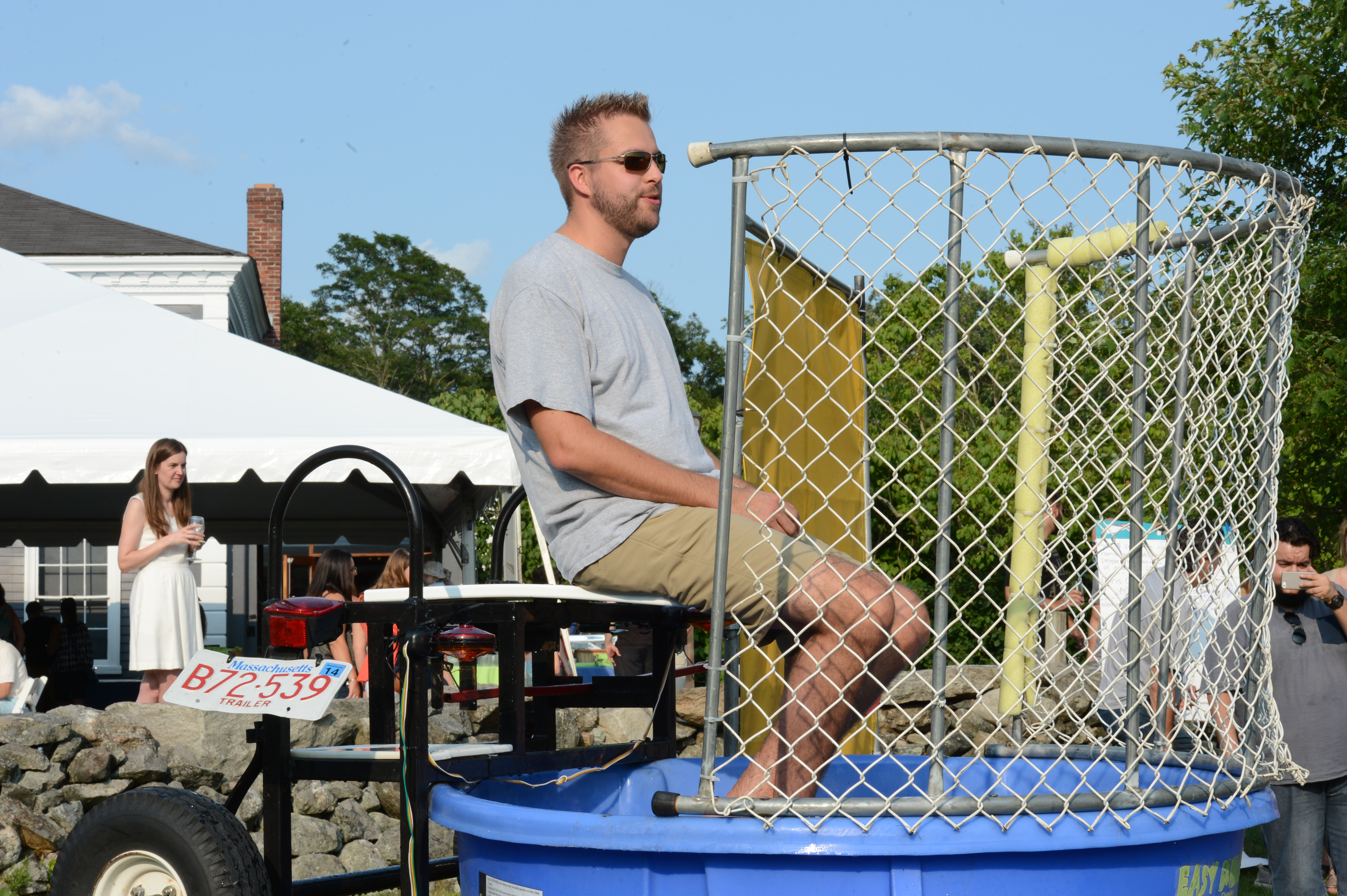 A man sits on a dunk tank platform, waiting to be dropped into the water at an outdoor event.