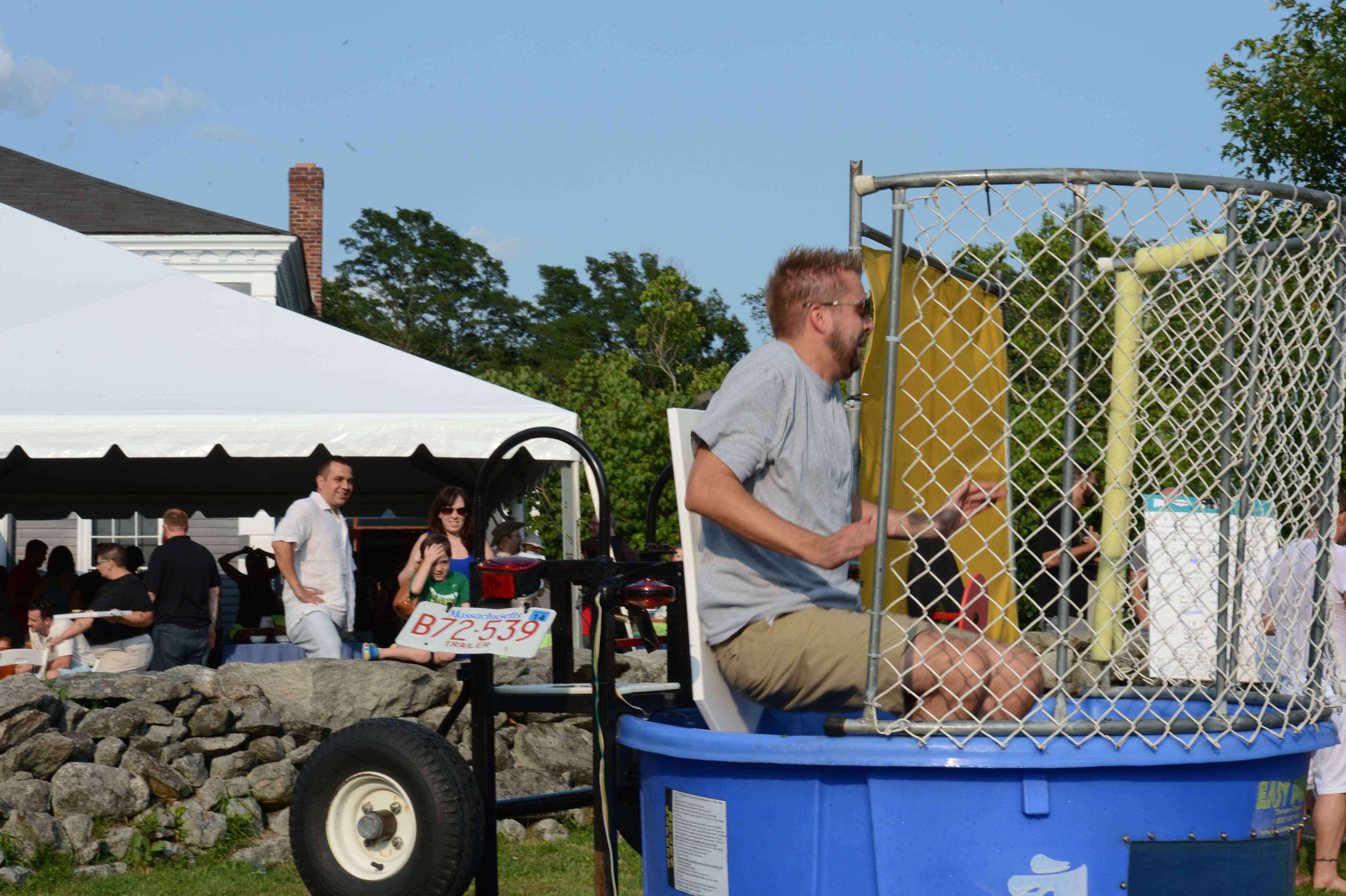 A man sits on a dunk tank seat, preparing to be dropped into the water at an outdoor event.