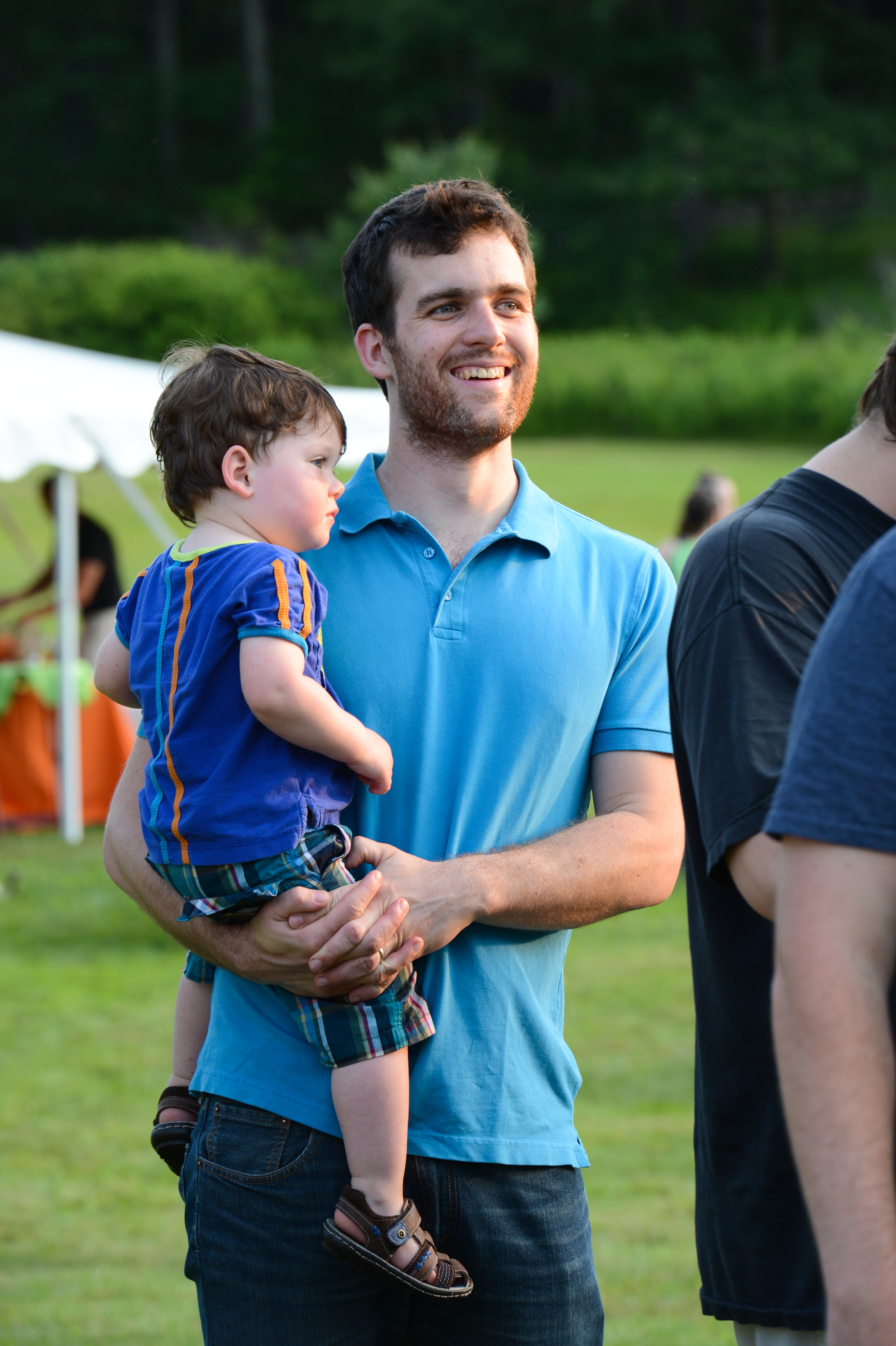 A man in a blue shirt holds a young child while watching an event at an outdoor gathering.