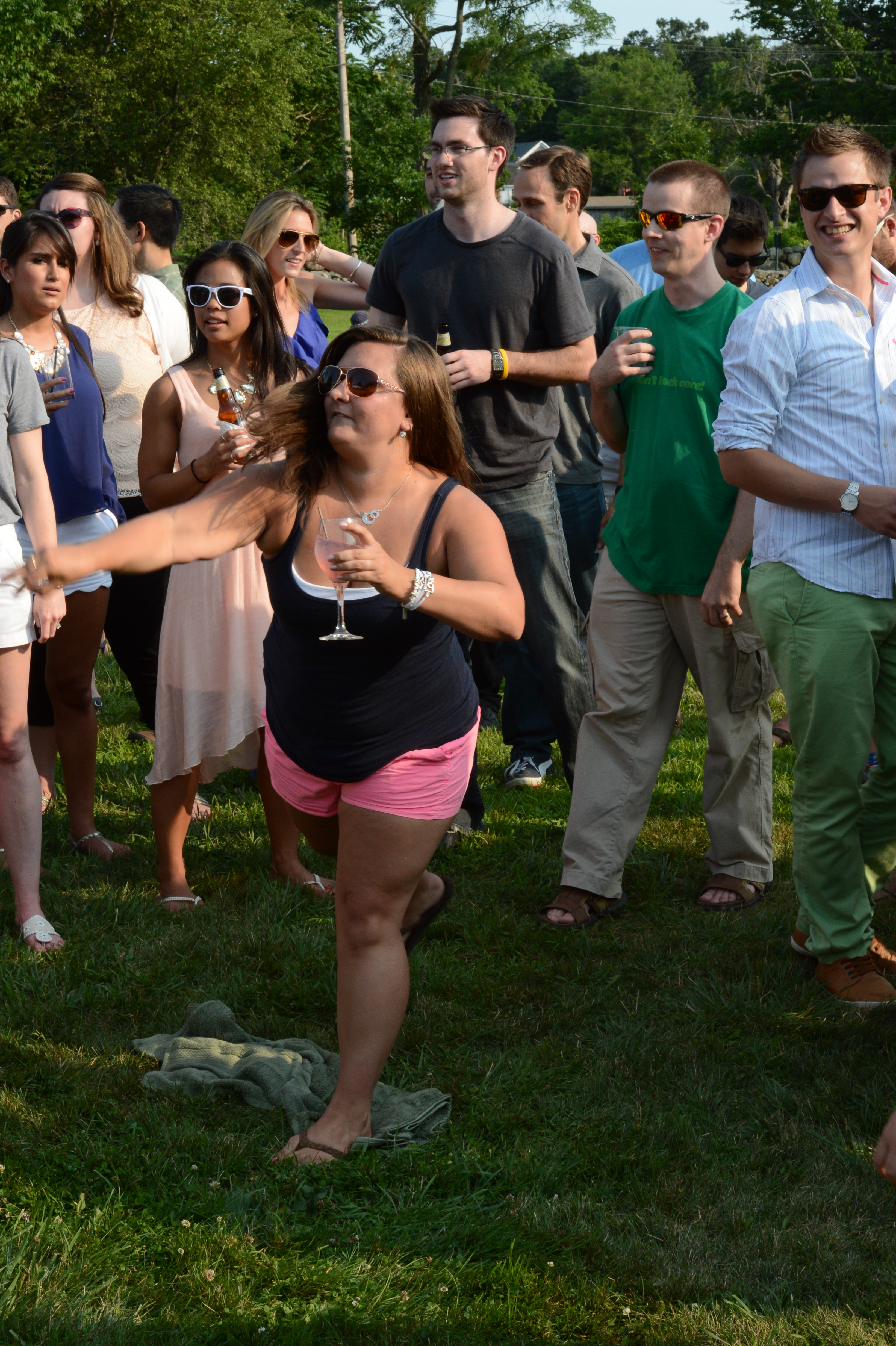 A woman throws a ball at a dunk tank target while holding a drink, surrounded by a cheering crowd.