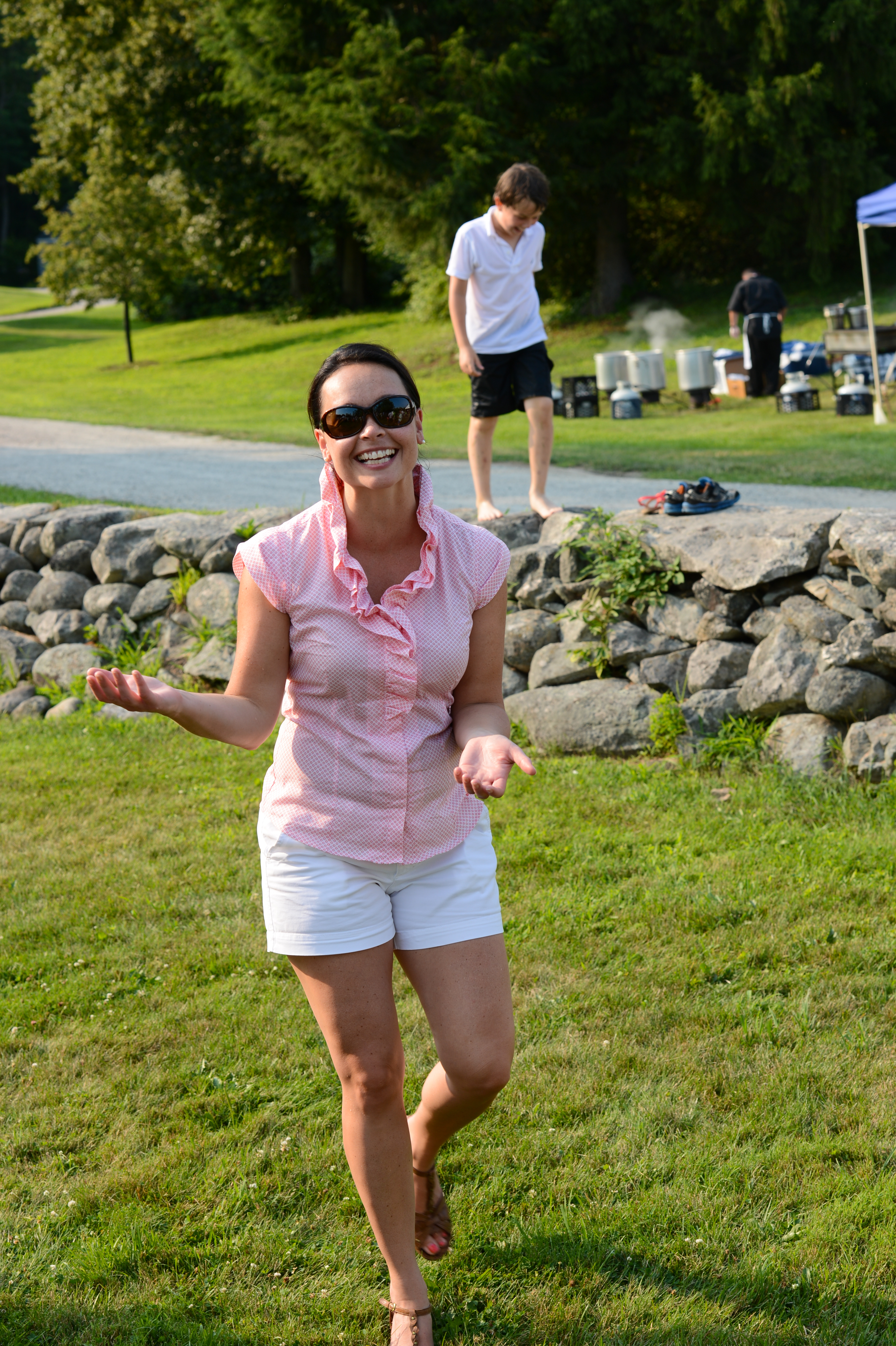 A smiling woman in sunglasses walks on the grass, gesturing with her hands at an outdoor summer gathering.