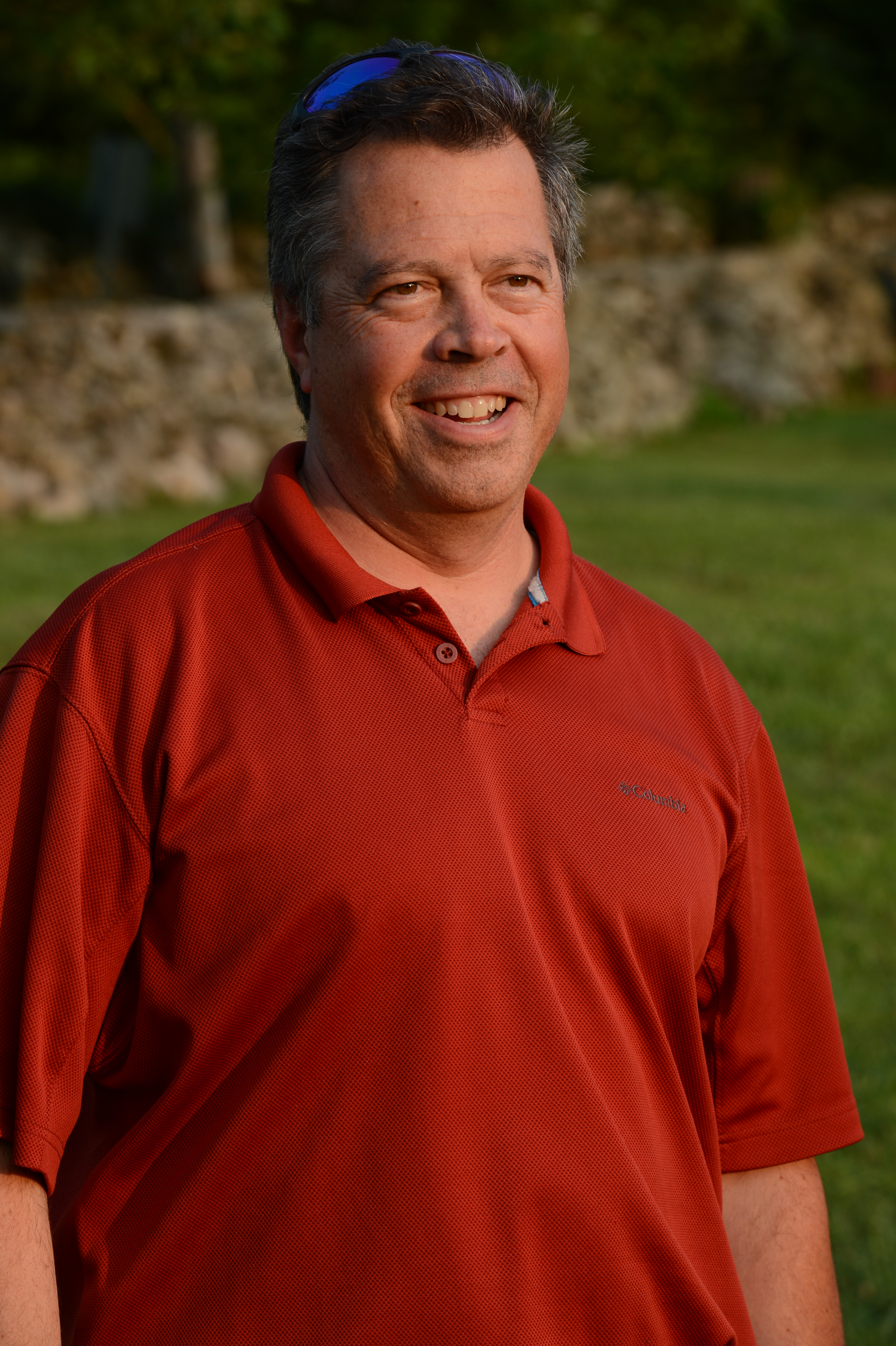 A man in a red polo shirt smiles while standing outdoors at a summer party.