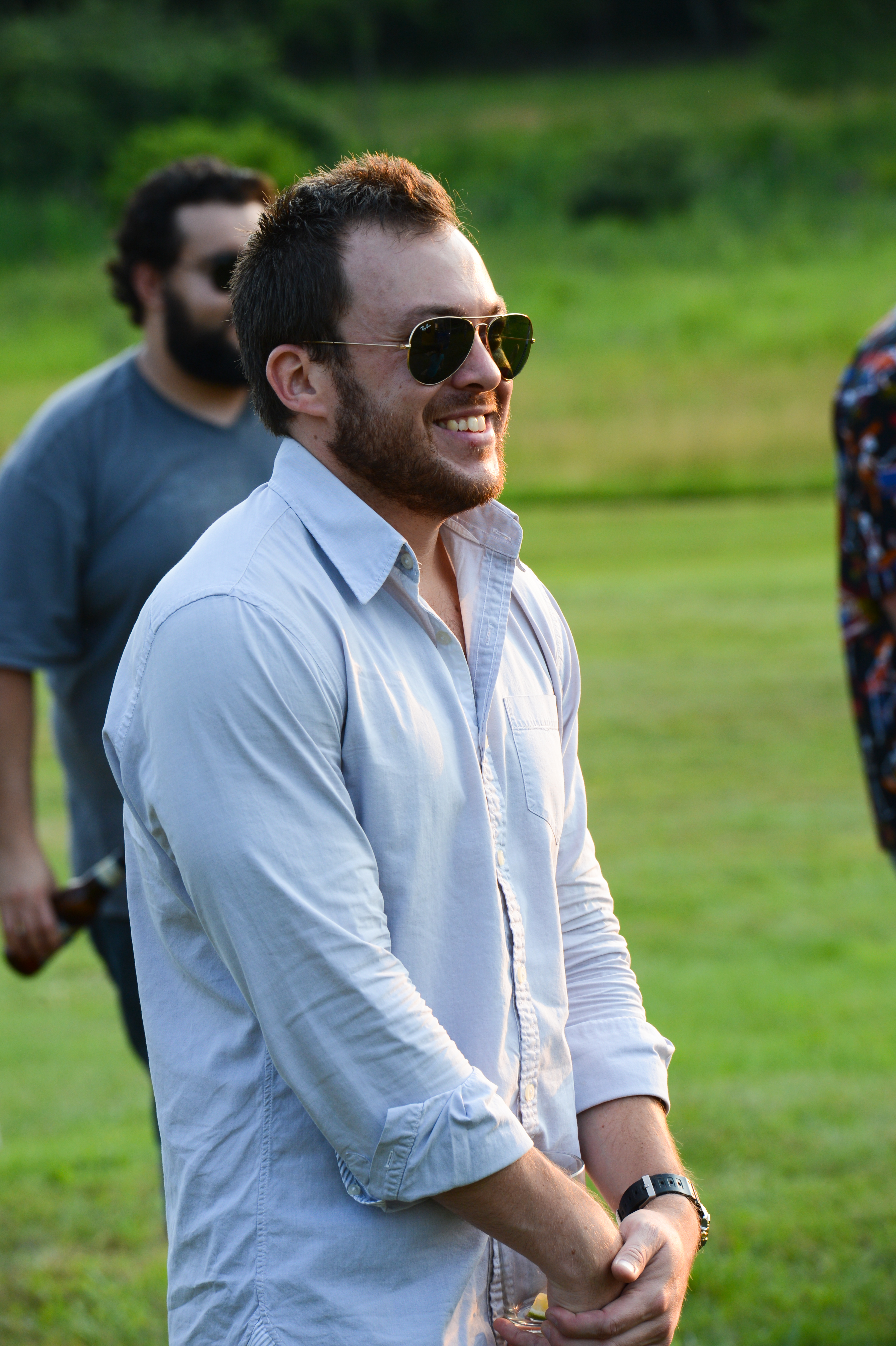 A man in sunglasses and a light shirt smiles while standing outdoors at a summer party.