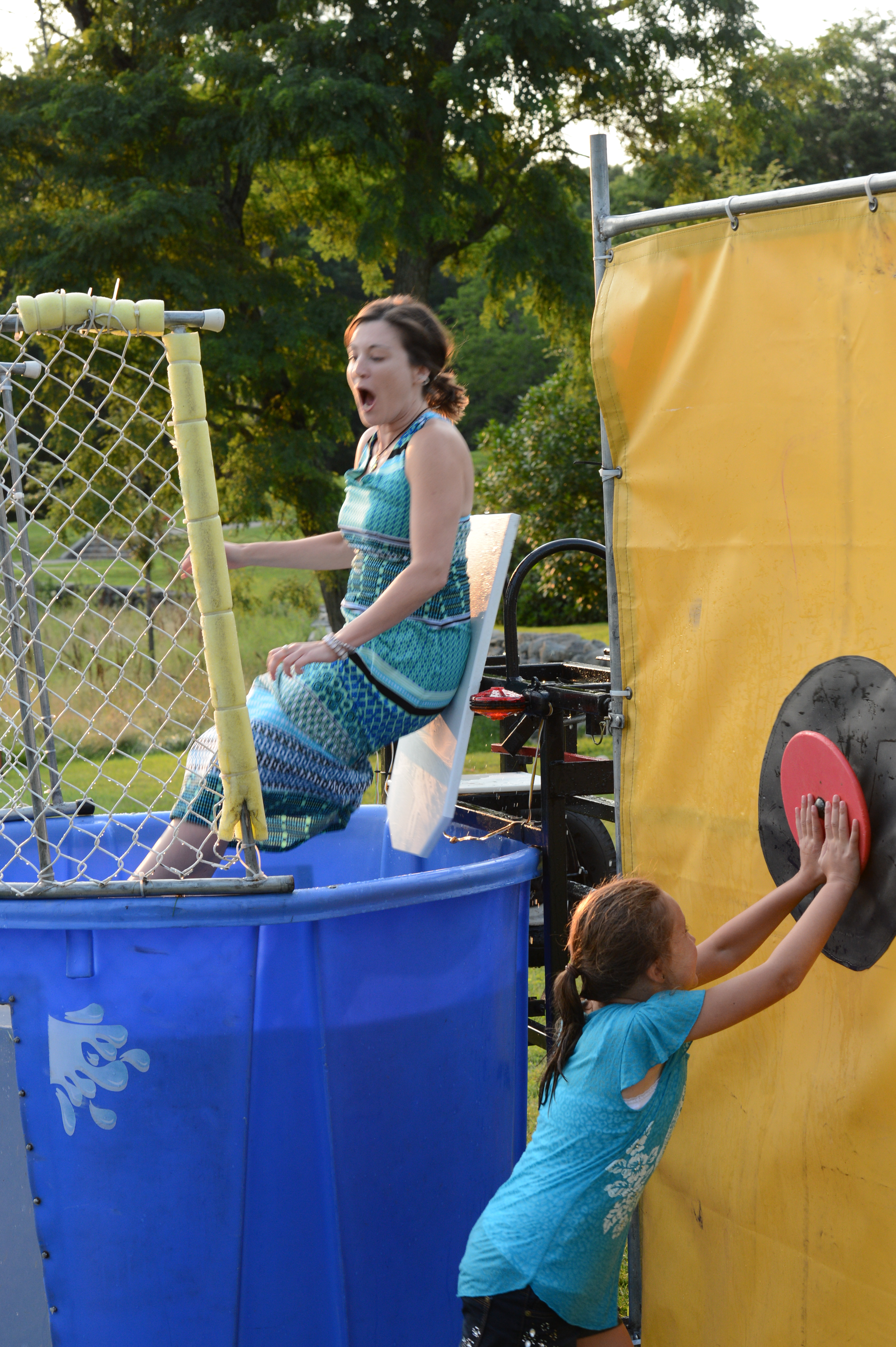 A young girl presses a button, triggering a woman to fall into a dunk tank at a summer party.