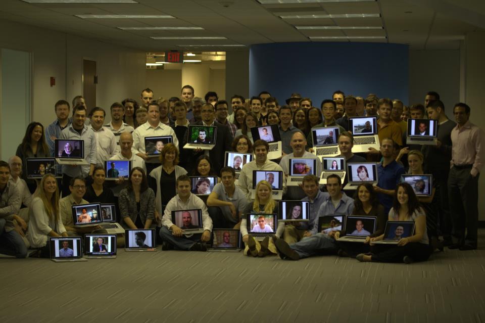 A large group of people poses together, holding laptops showing images of remote participants.