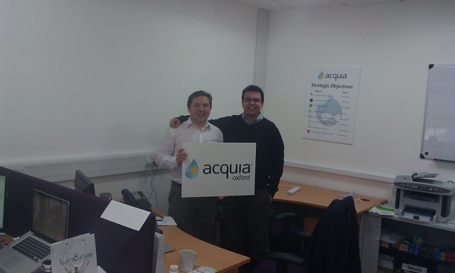 Two men stand in an office, smiling and holding an "Acquia Oxford" sign.