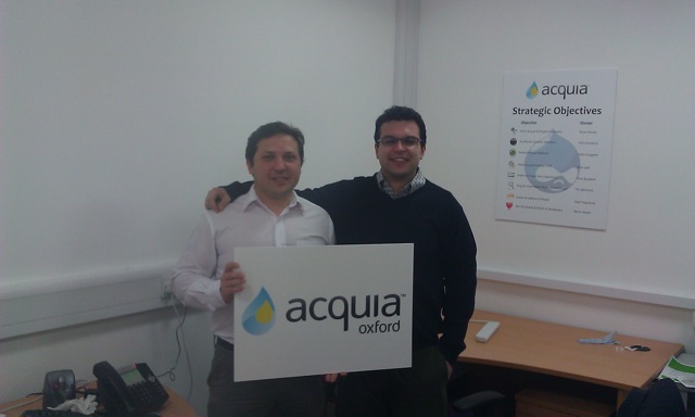 Two men stand in an office, smiling and holding a sign that reads "Acquia Oxford.