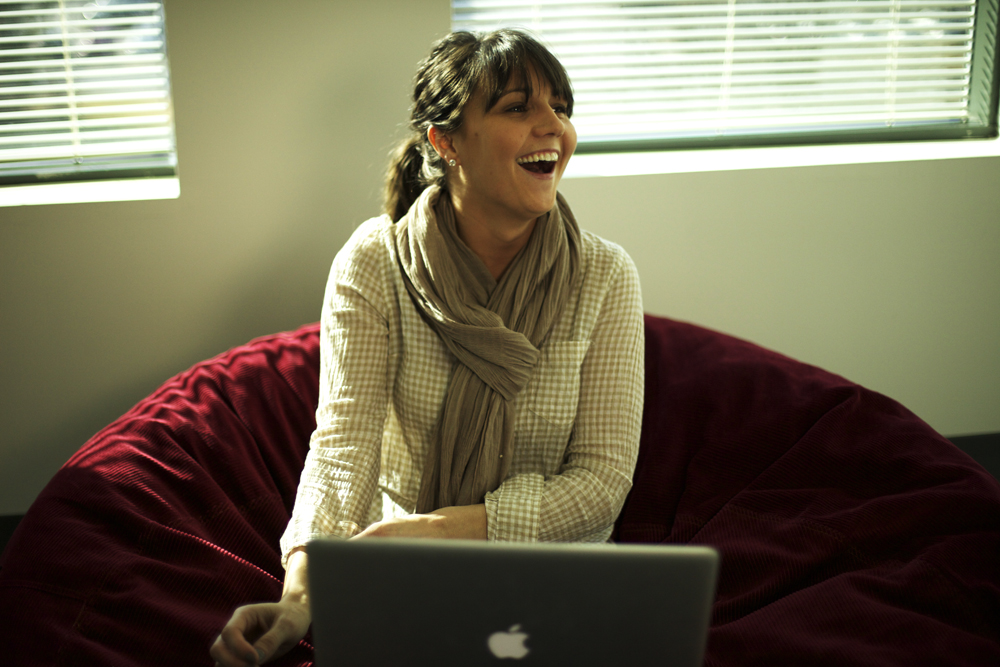 A woman sits on a bean bag, smiling and laughing while using a laptop.