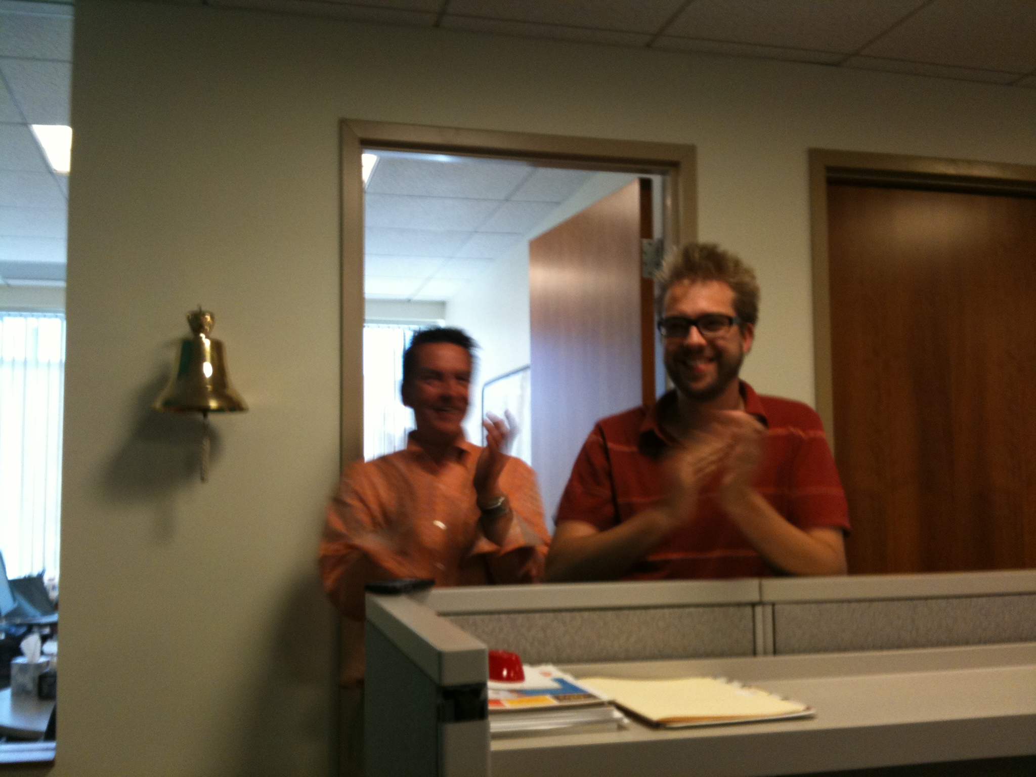 Two people stand near a wall-mounted bell, clapping and smiling in an office setting.