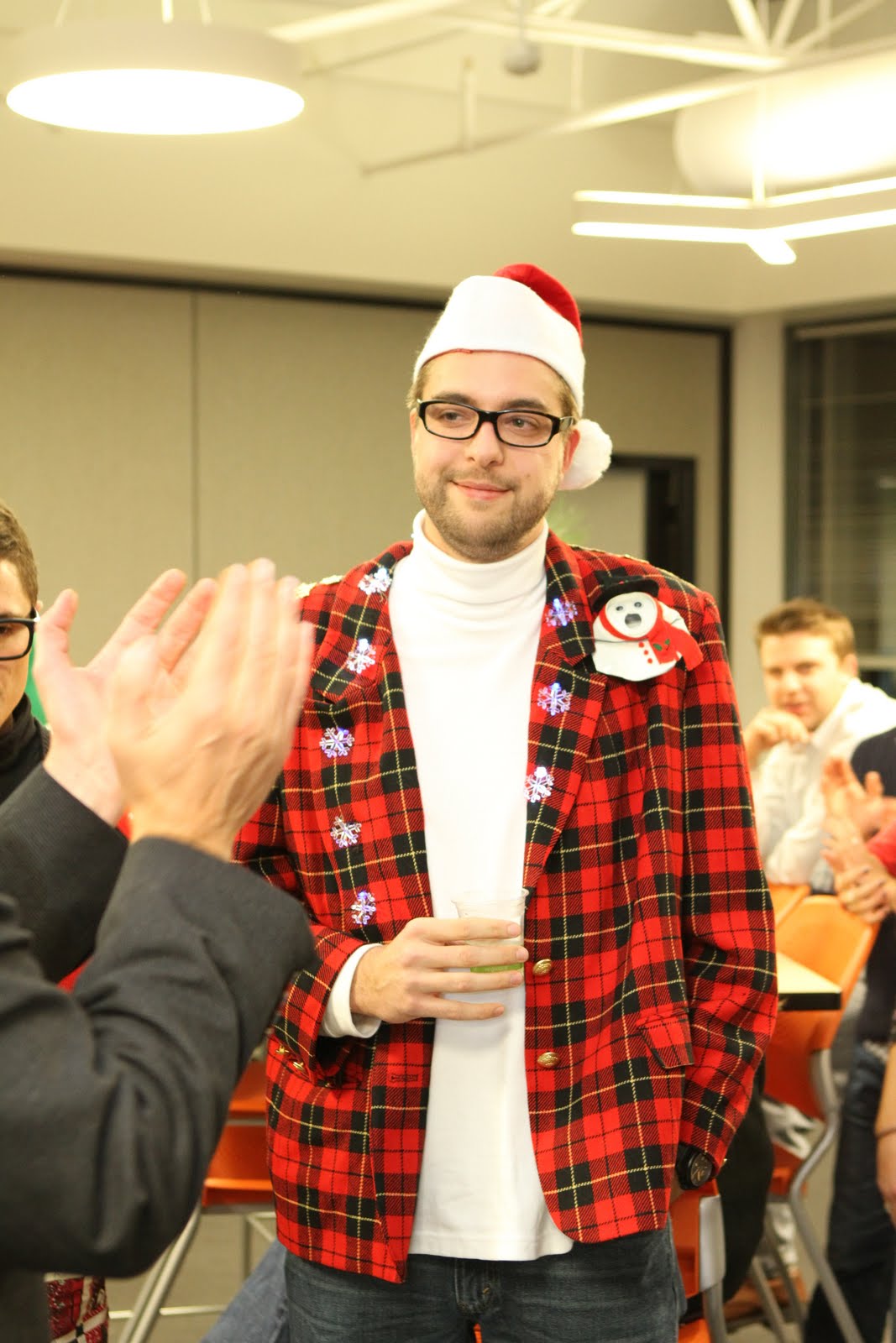 A man in a red plaid jacket and Santa hat holds a drink while people clap around him.