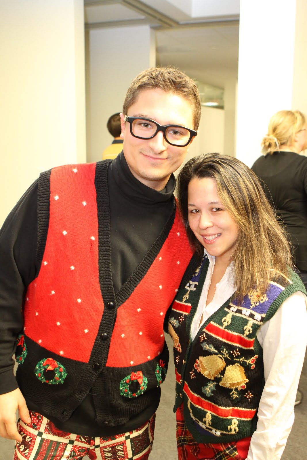 Two people wearing festive Christmas sweaters smile at the camera during a holiday party.