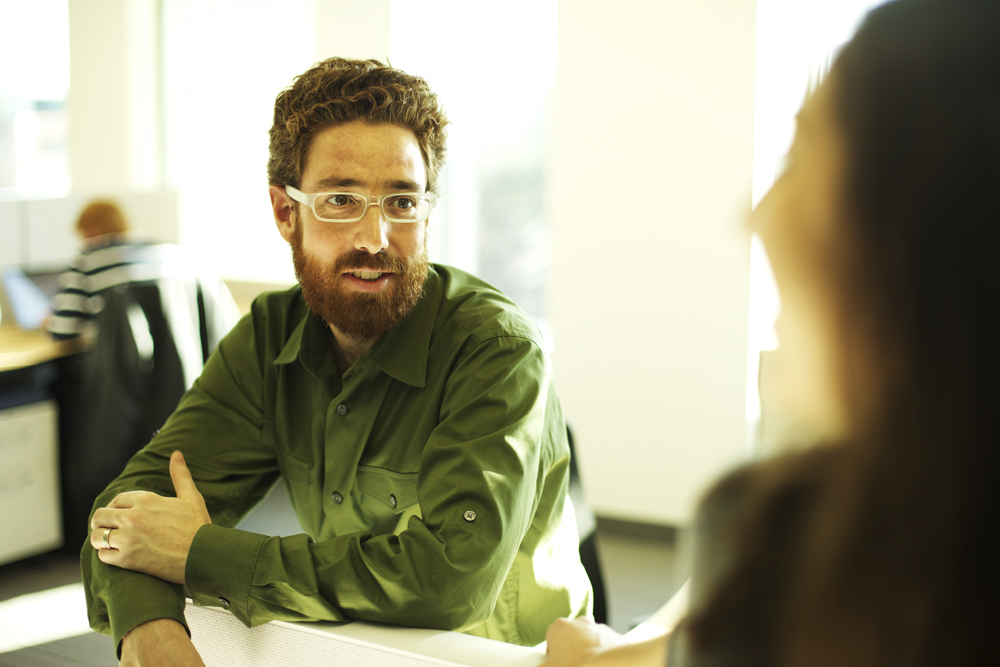 A man in a green shirt and glasses sits and talks with a woman in an office.