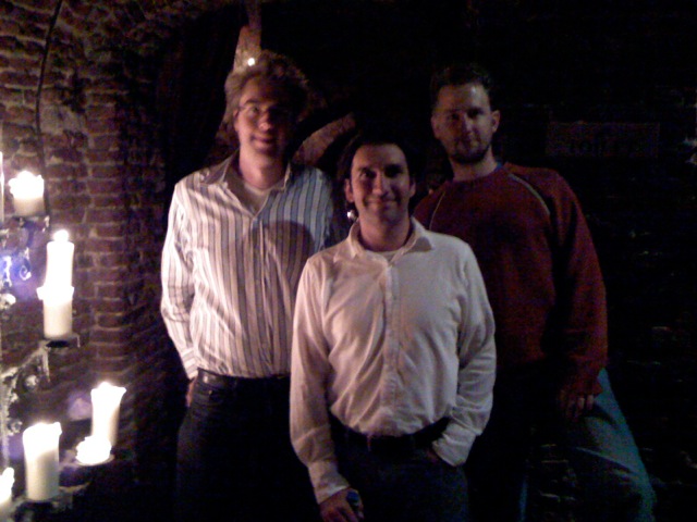 Three men stand together at the first Acquia company meeting in a dimly lit room with brick walls and candles.