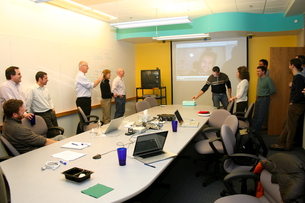 A group of people gathers in a conference room to celebrate Drupal's eighth birthday with a cake and video call.