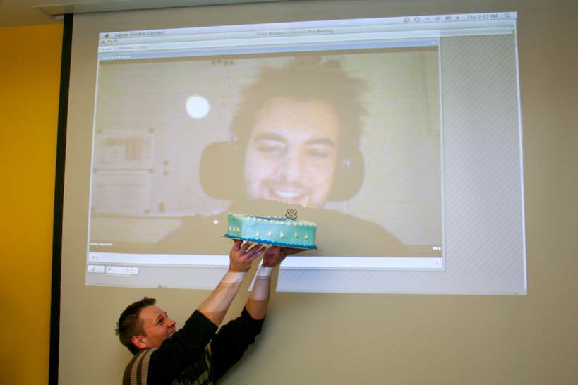 A person holds up a blue birthday cake with an "8" topper in front of a video projection of a smiling person.