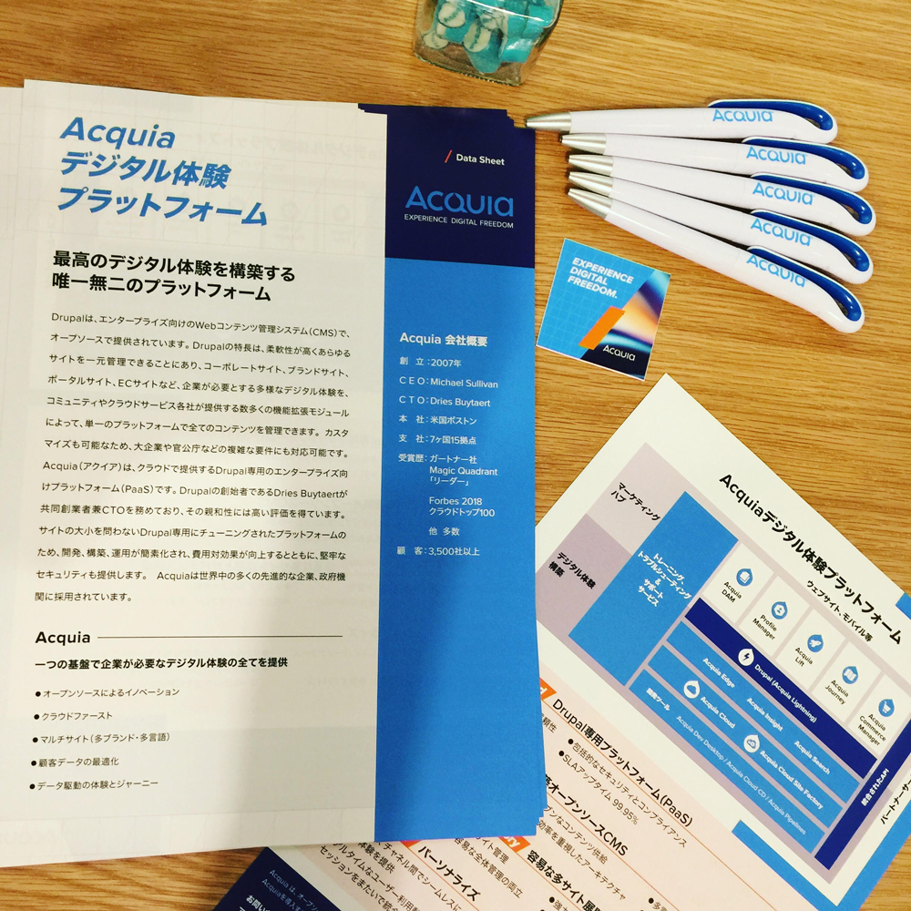 A Japanese-language Acquia brochure and marketing materials are neatly arranged on a wooden table with branded pens.