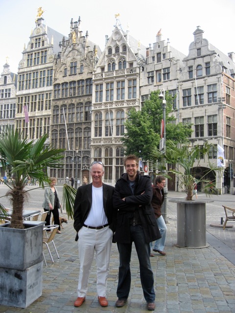 Two men stand smiling in a European city square.