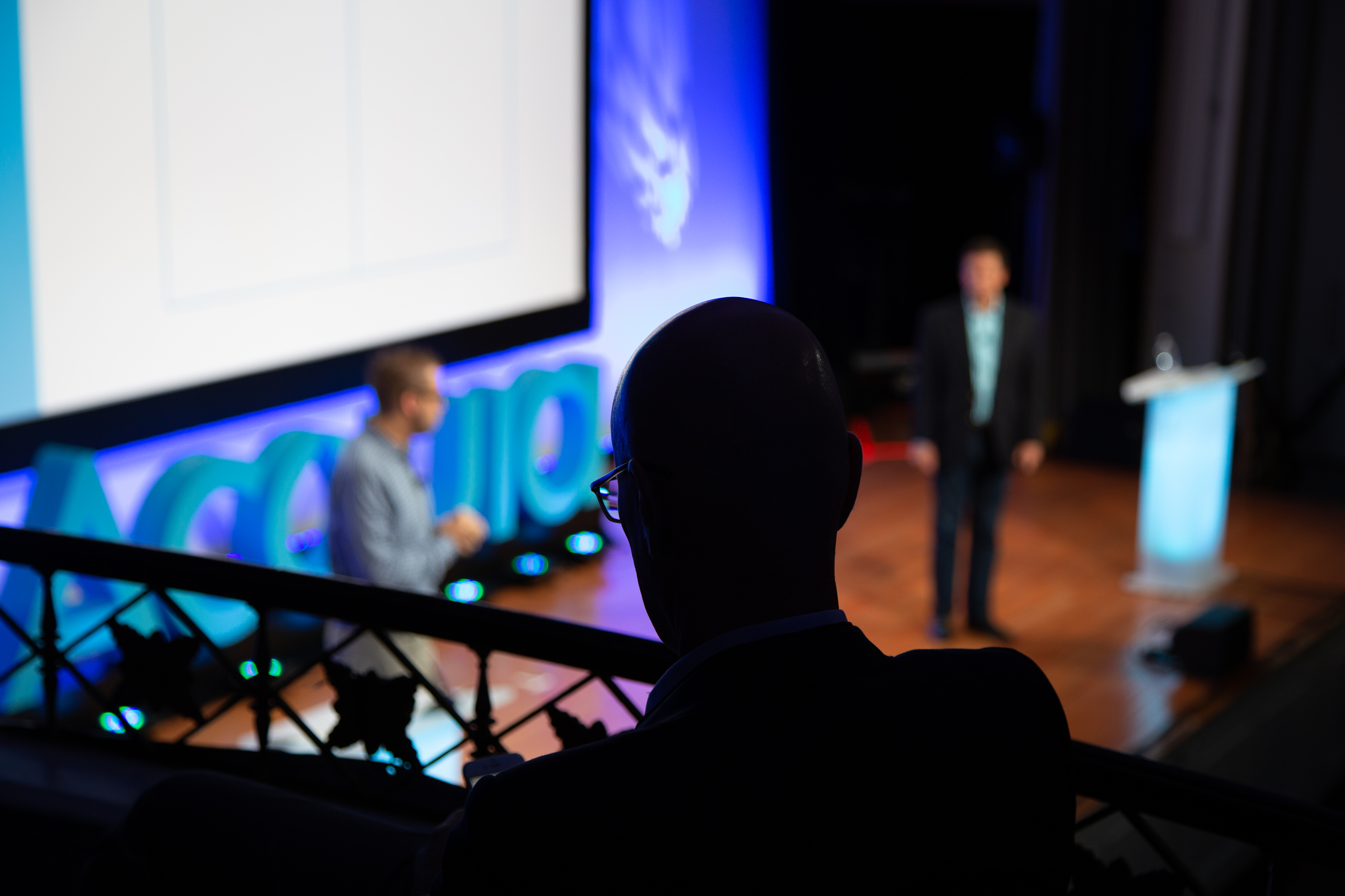 An audience member watches two speakers on stage at a conference with an "Acquia" sign in the background.