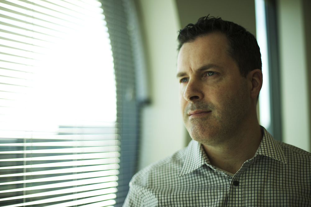 A man in a checkered shirt looks out of a window with blinds, appearing thoughtful.