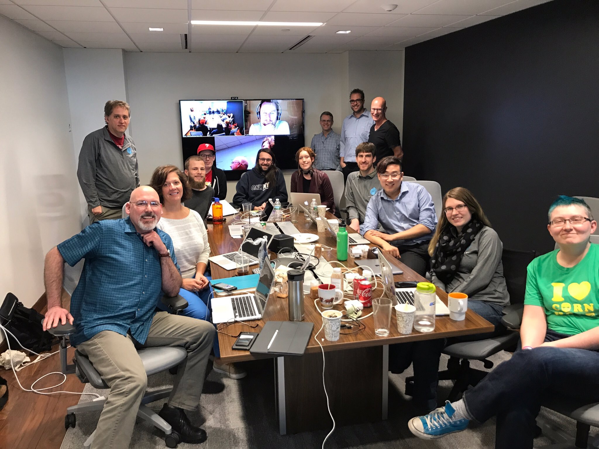 A group of people in a conference room sitting around a table with laptops, smiling at the camera.
