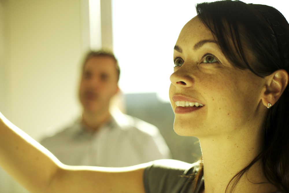 A woman smiles while writing on a whiteboard, with a man observing in the background.