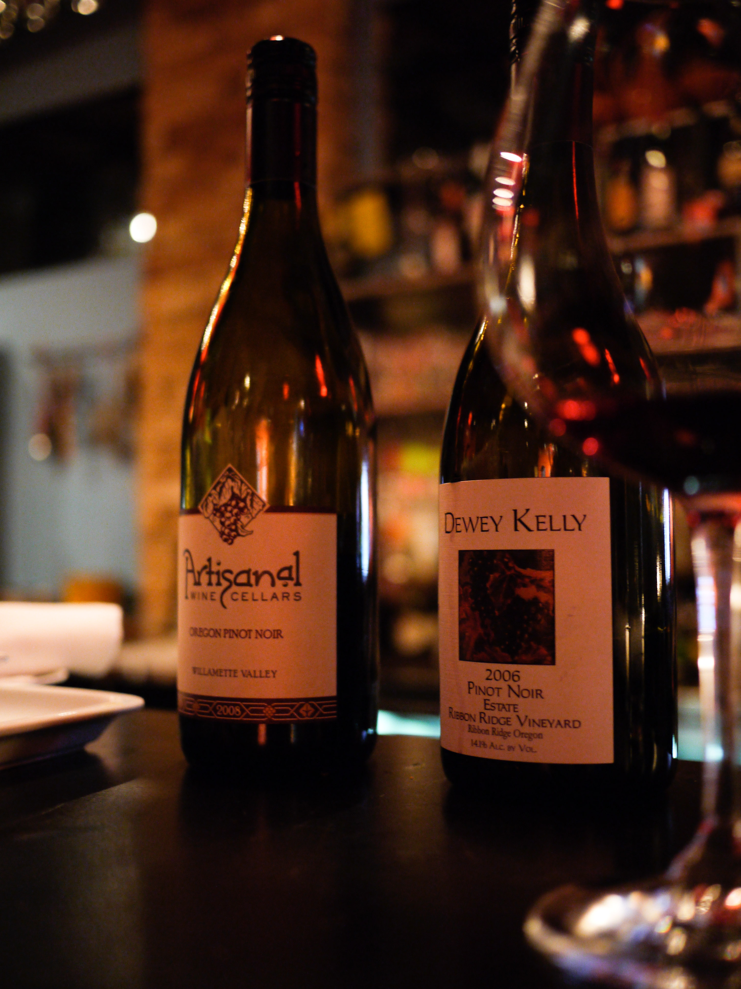 Two bottles of Pinot Noir wine on a bar counter, with a glass of red wine partially visible.