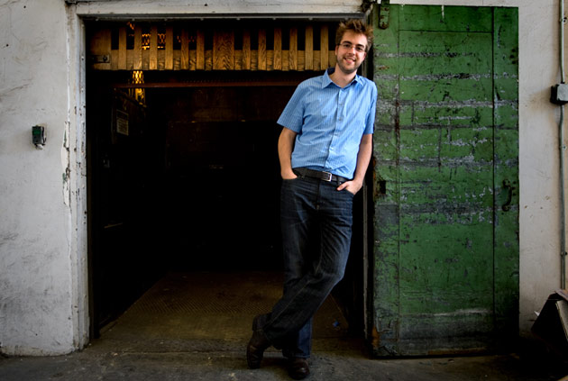 A man in a blue shirt leans against a doorway, smiling at the camera.