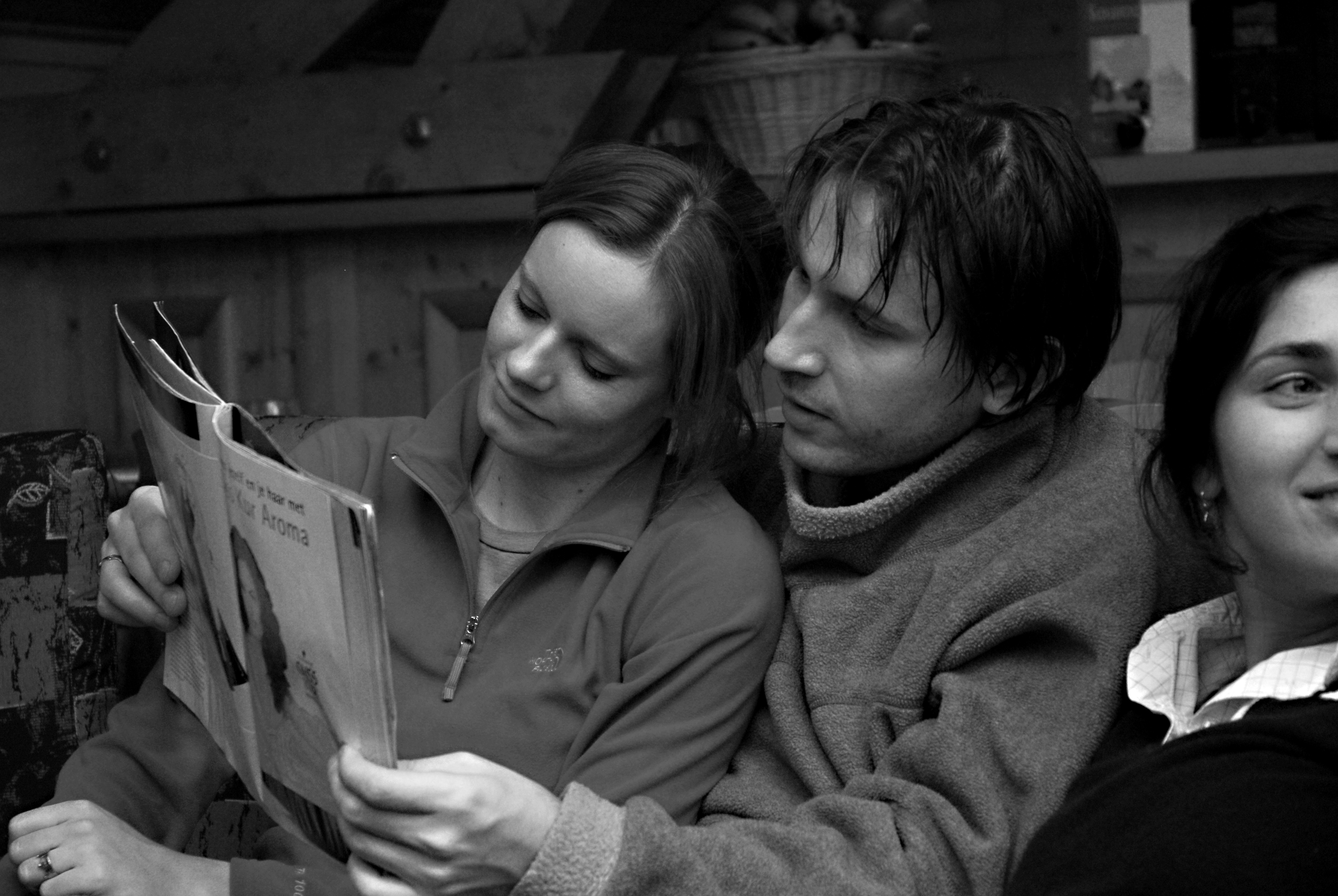 A man and woman sit closely together on a couch, reading a magazine, while another woman smiles nearby.