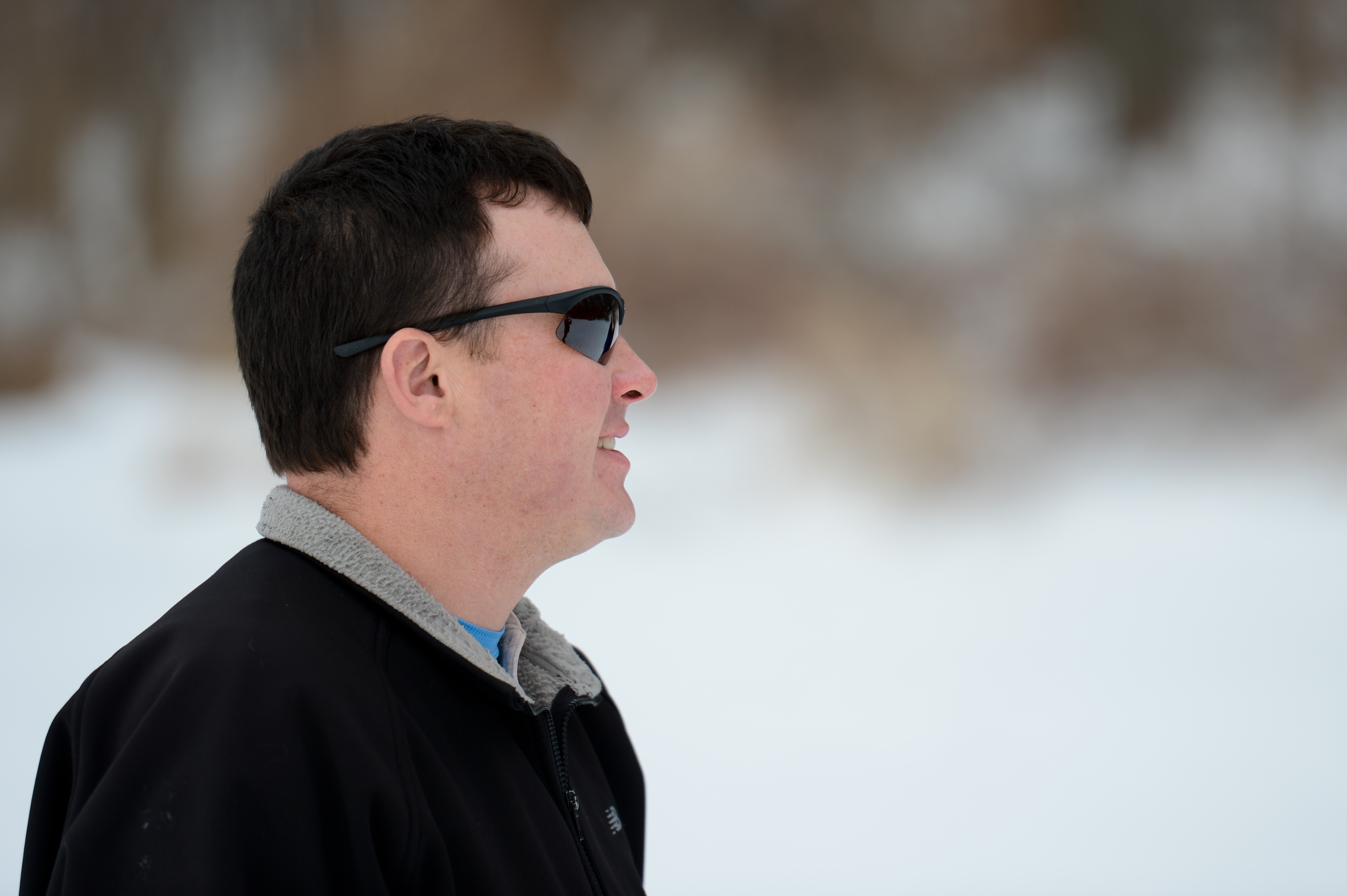 A man wearing sunglasses and a black jacket looks to the side in a snowy outdoor setting.