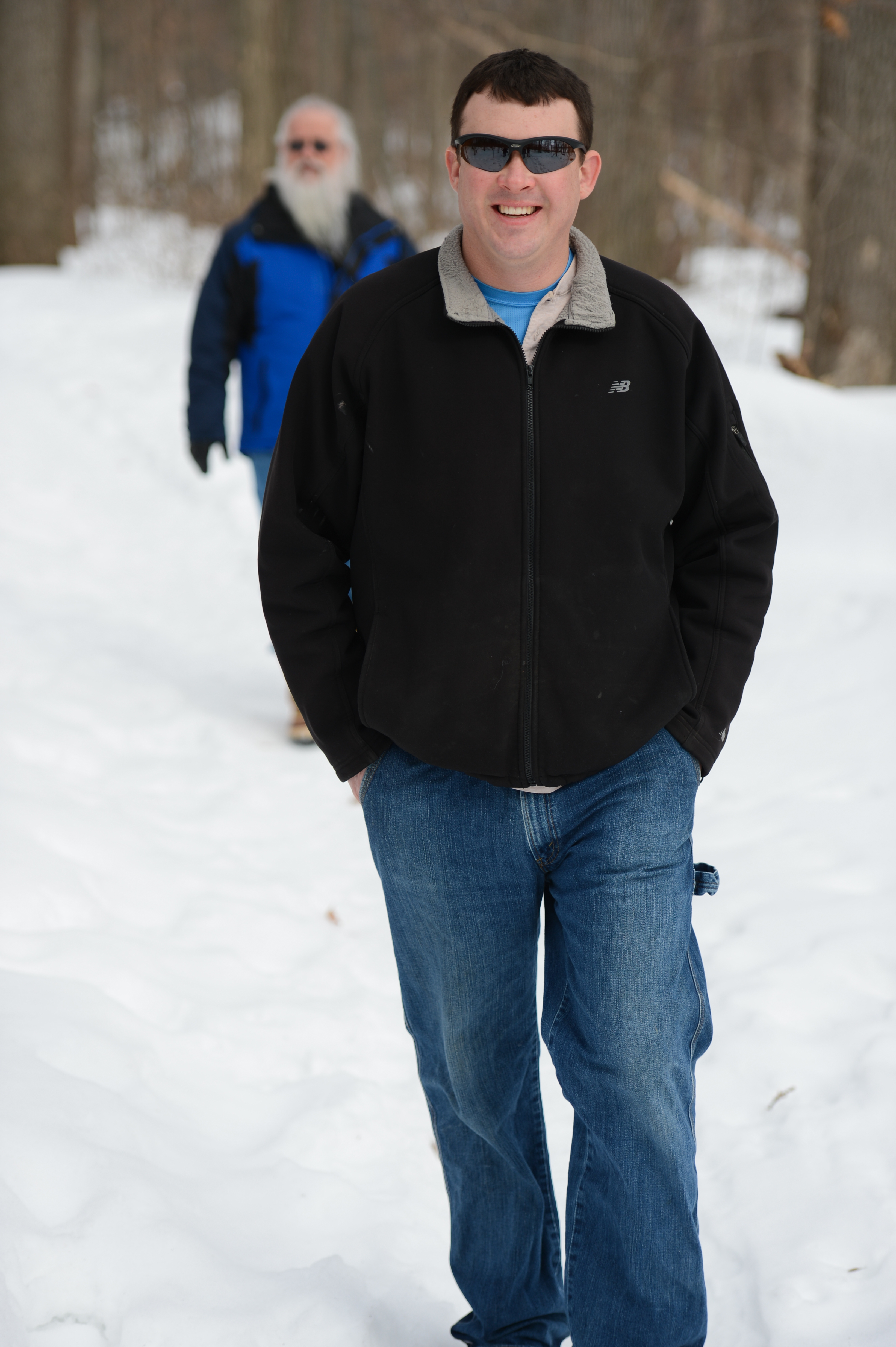 A man in sunglasses and a black jacket walks on a snowy path, with another person following behind.