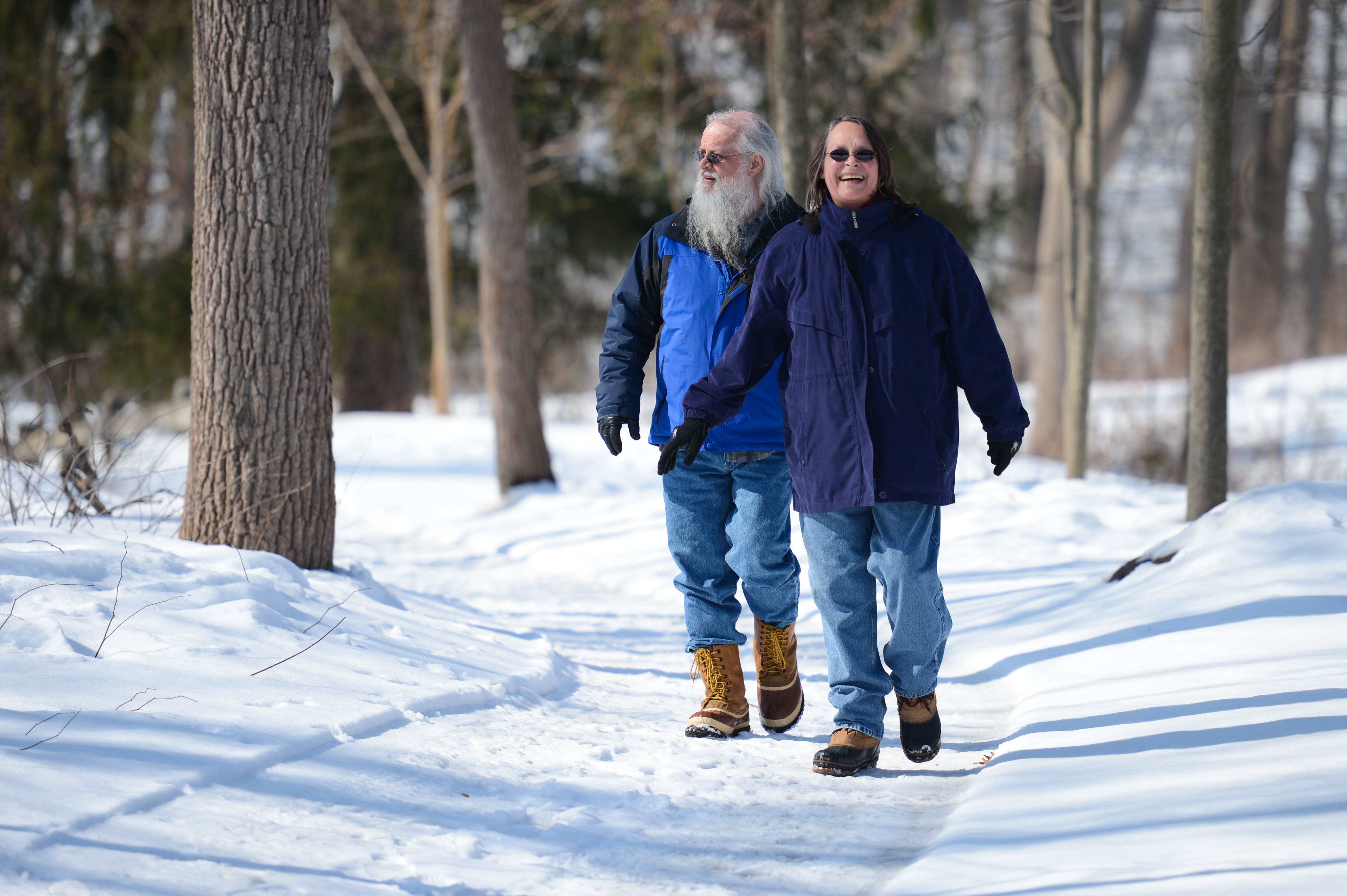 Two people wearing winter coats and boots walk along a snowy path in a forested area.