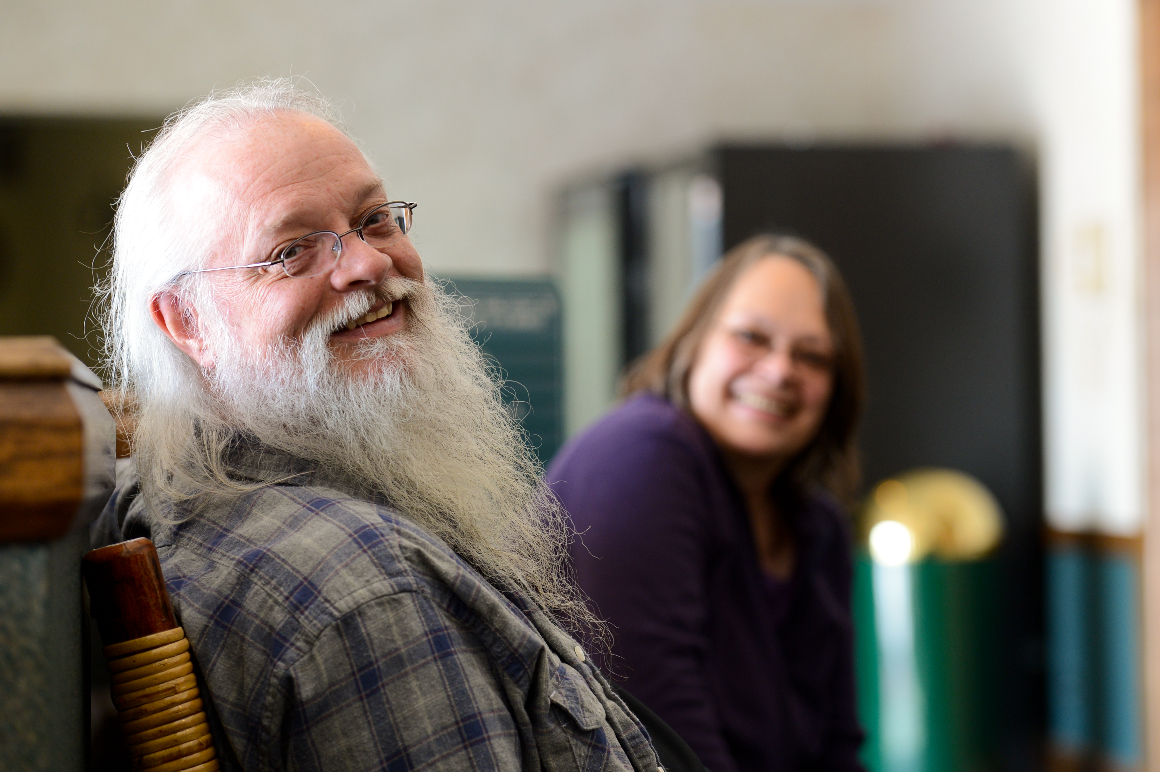 A bearded man in glasses smiles while sitting on a chair, with a woman smiling in the background.