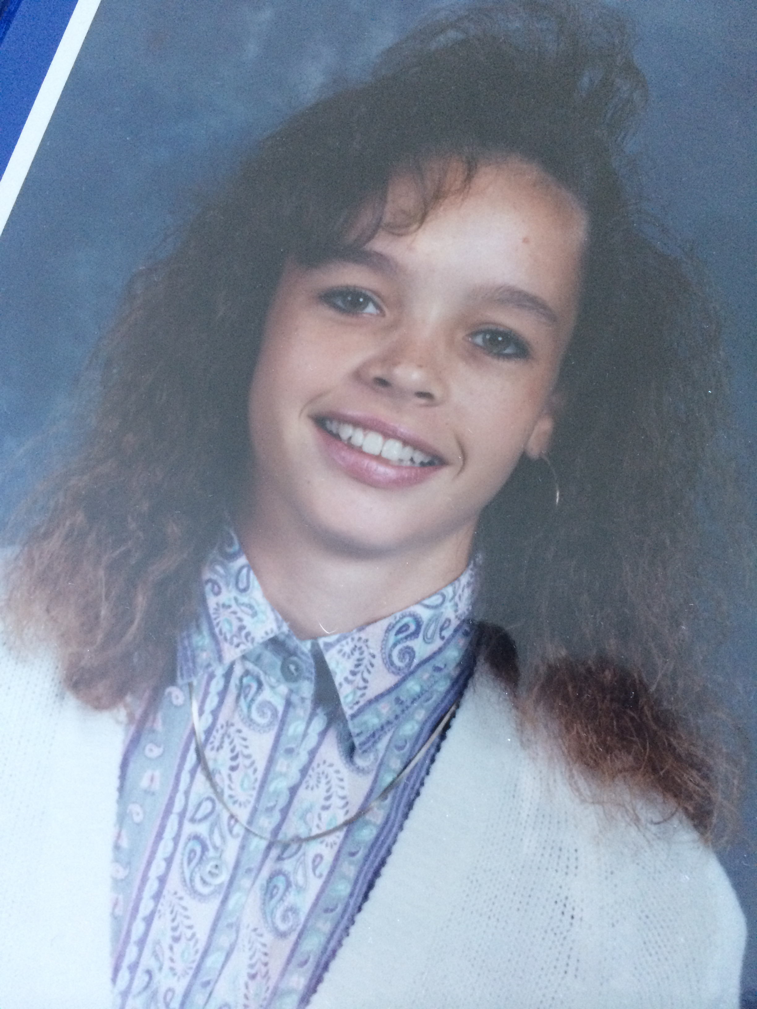 A person with curly hair smiles at the camera, wearing a patterned shirt, cardigan, and a necklace.