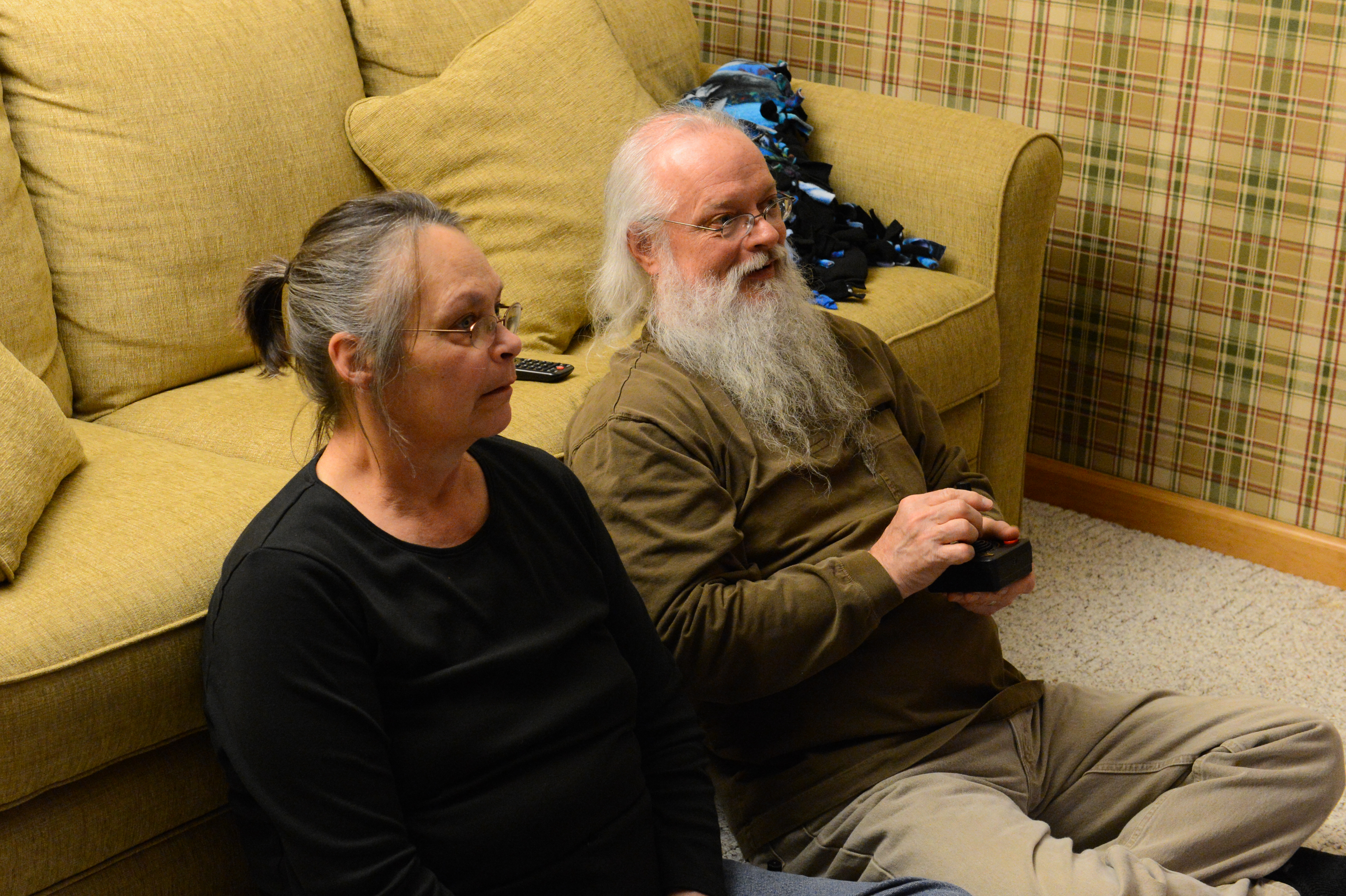 An older man and woman sit on the floor, watching something.