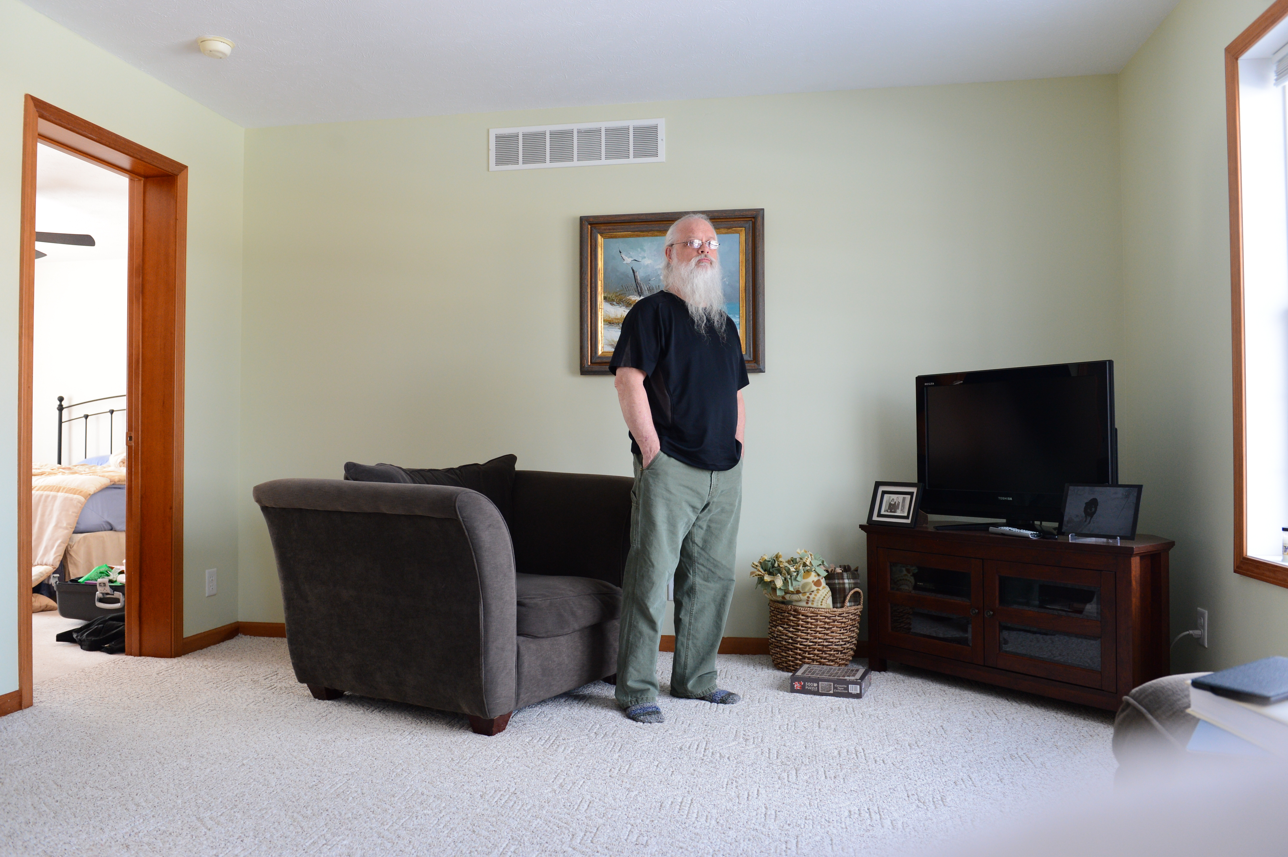 An older man with a long white beard stands in a living room near a couch, looking toward the camera.