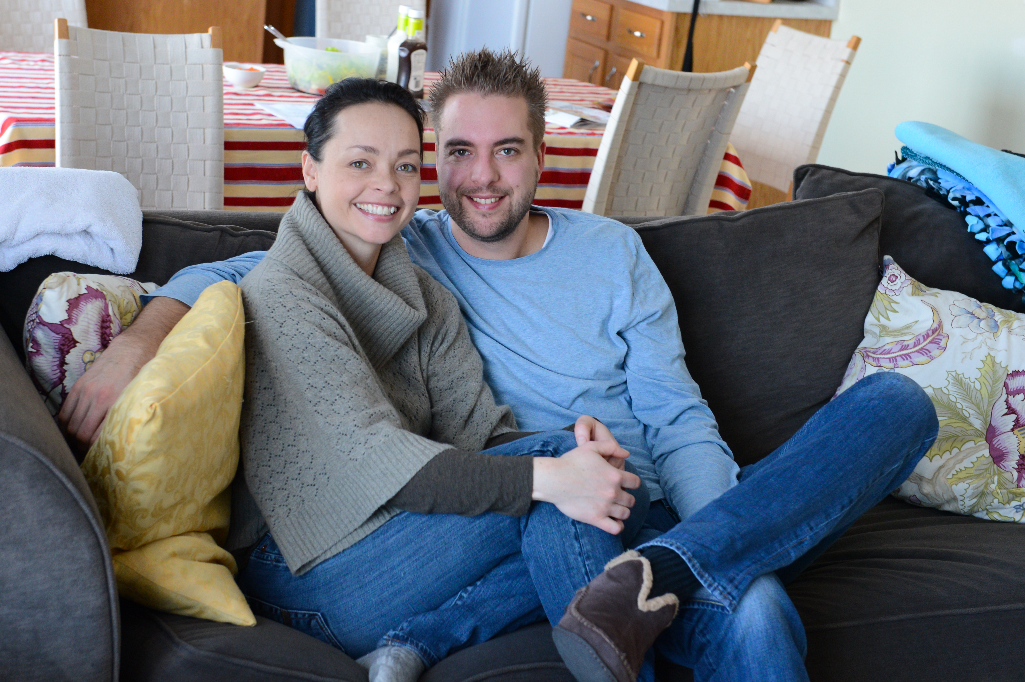 A smiling couple sits closely together on a couch, holding hands and looking at the camera.