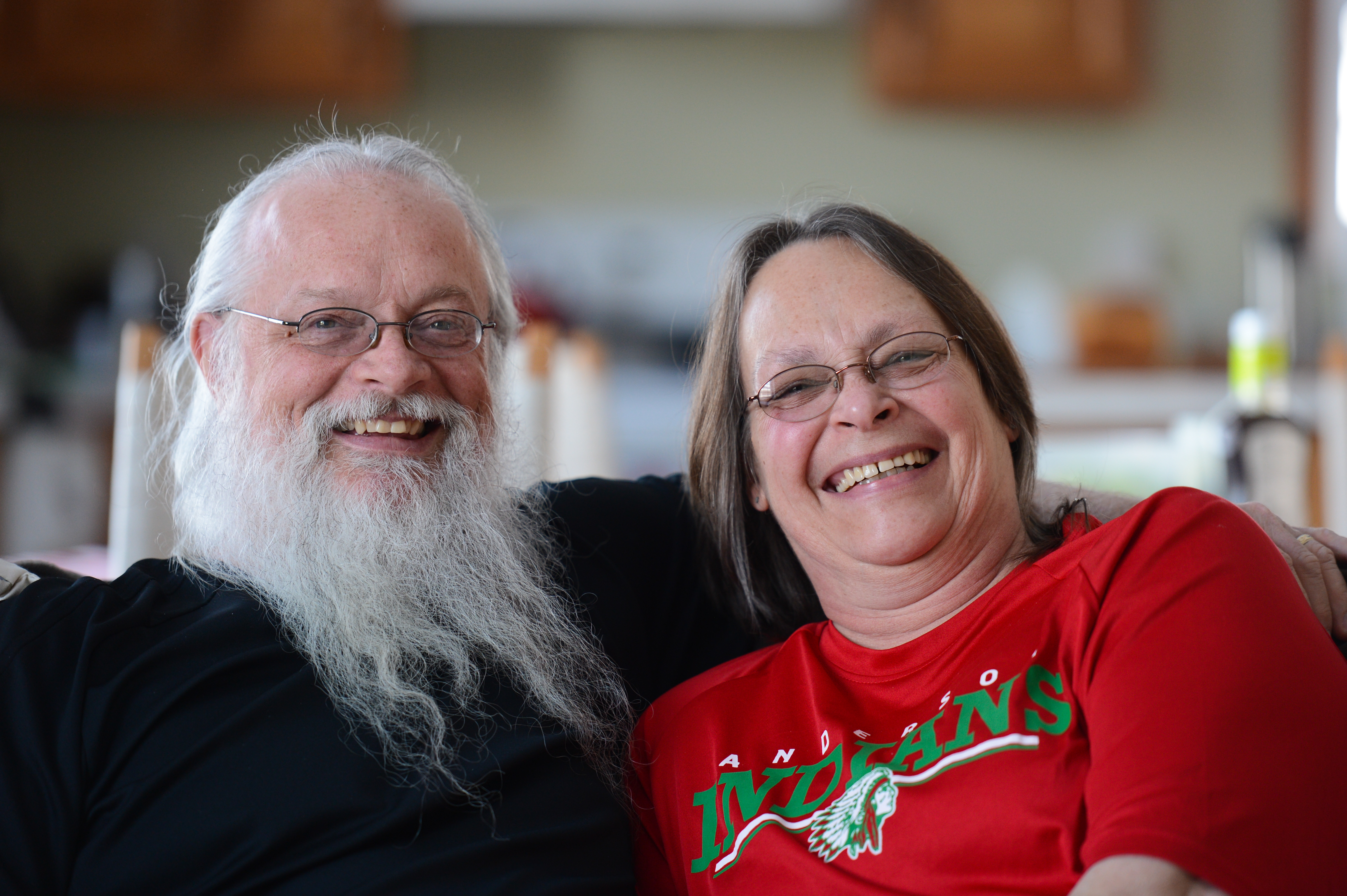 A smiling man and woman sit closely together, looking at the camera.