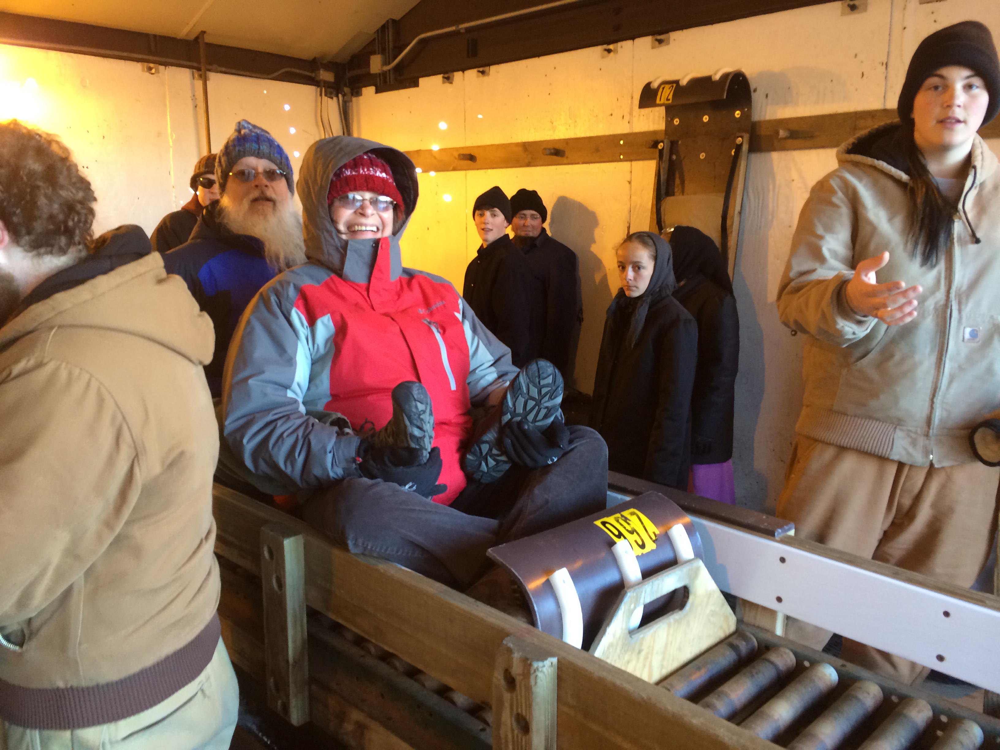 A person in a red and gray jacket sits on a wooden sled, smiling, while others stand nearby.