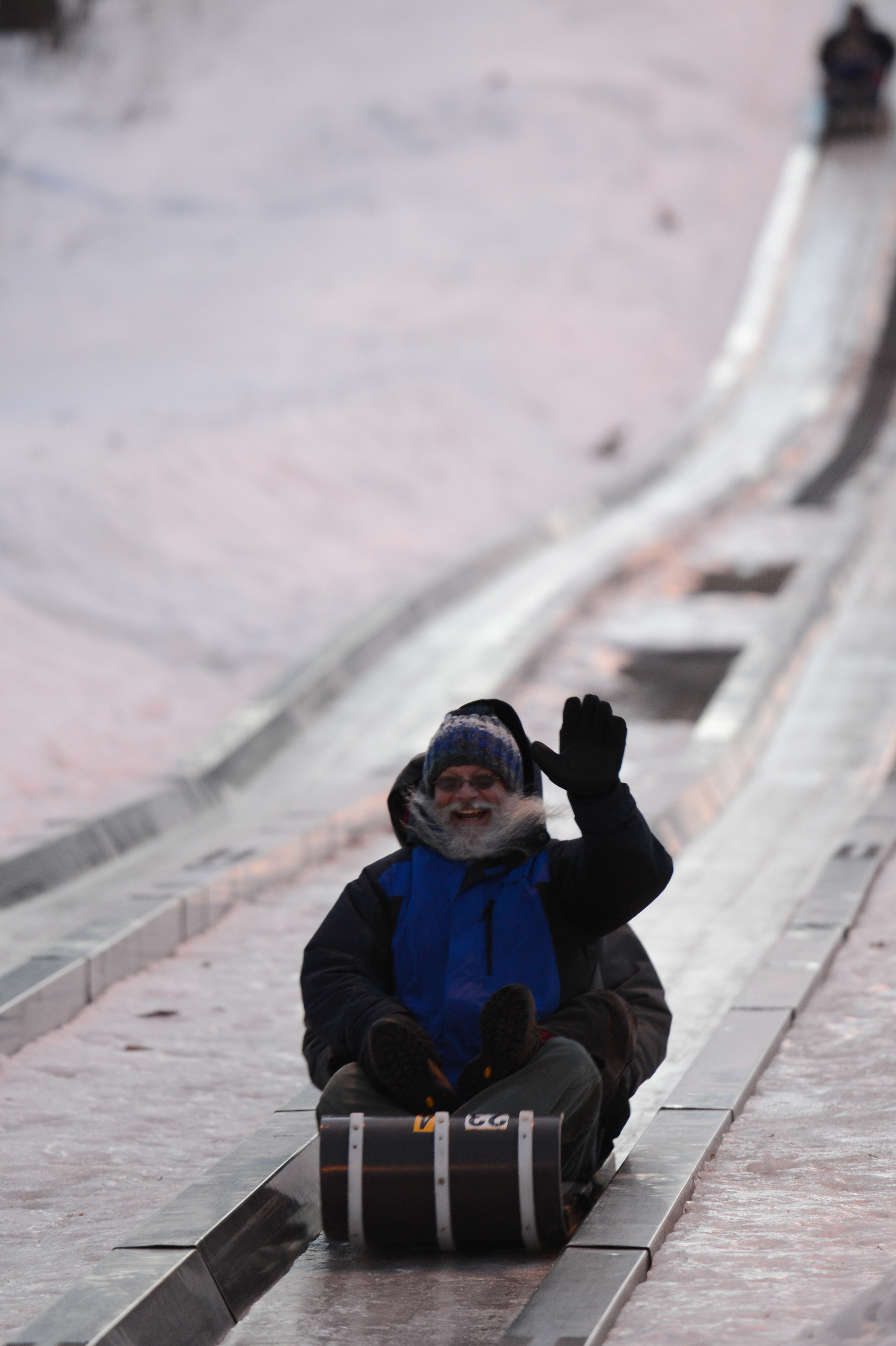 A person in winter clothing rides a sled down an icy slide, smiling and waving.