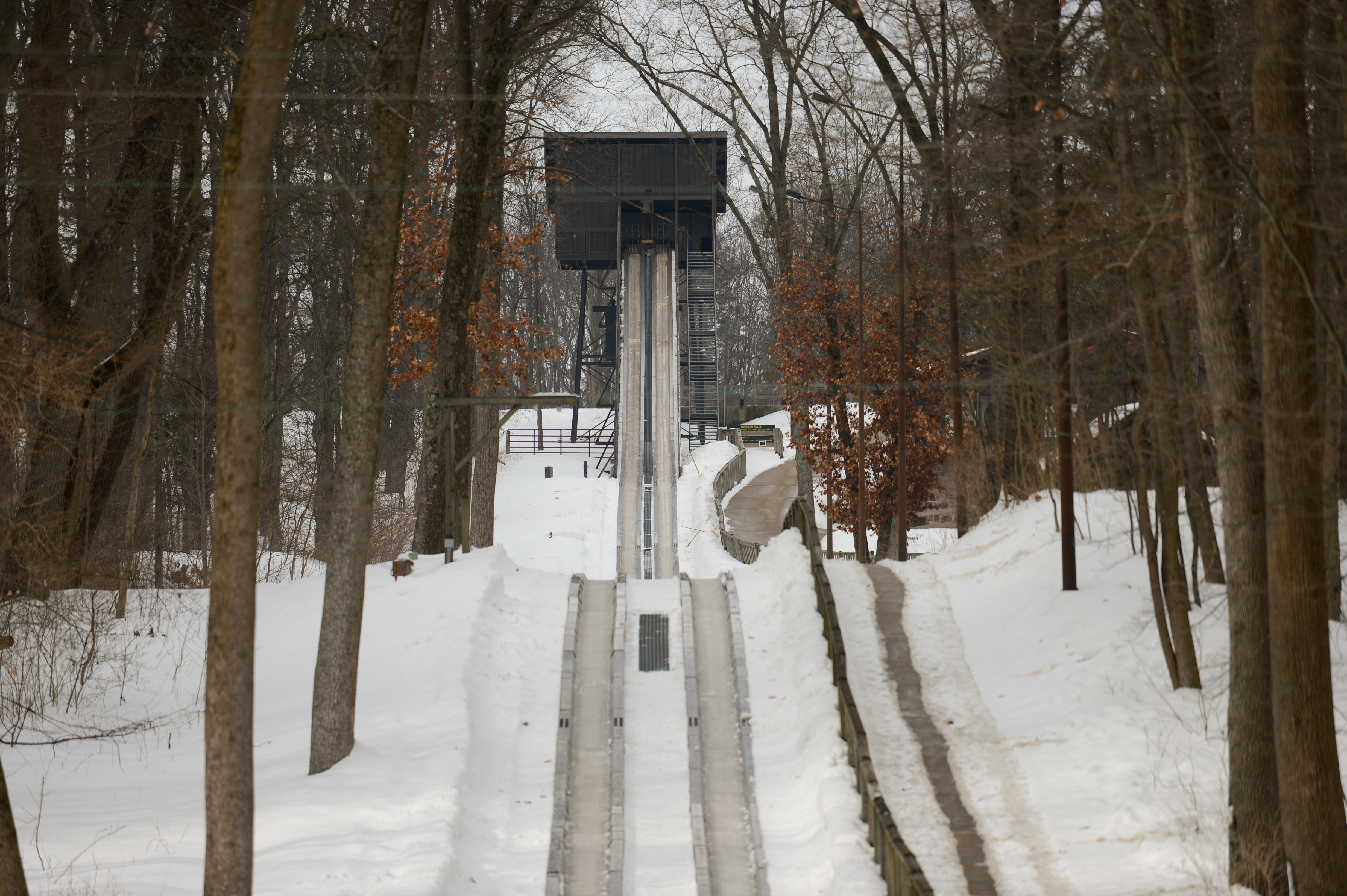 A snowy ski jump ramp surrounded by tall trees, leading up to a platform in the distance.