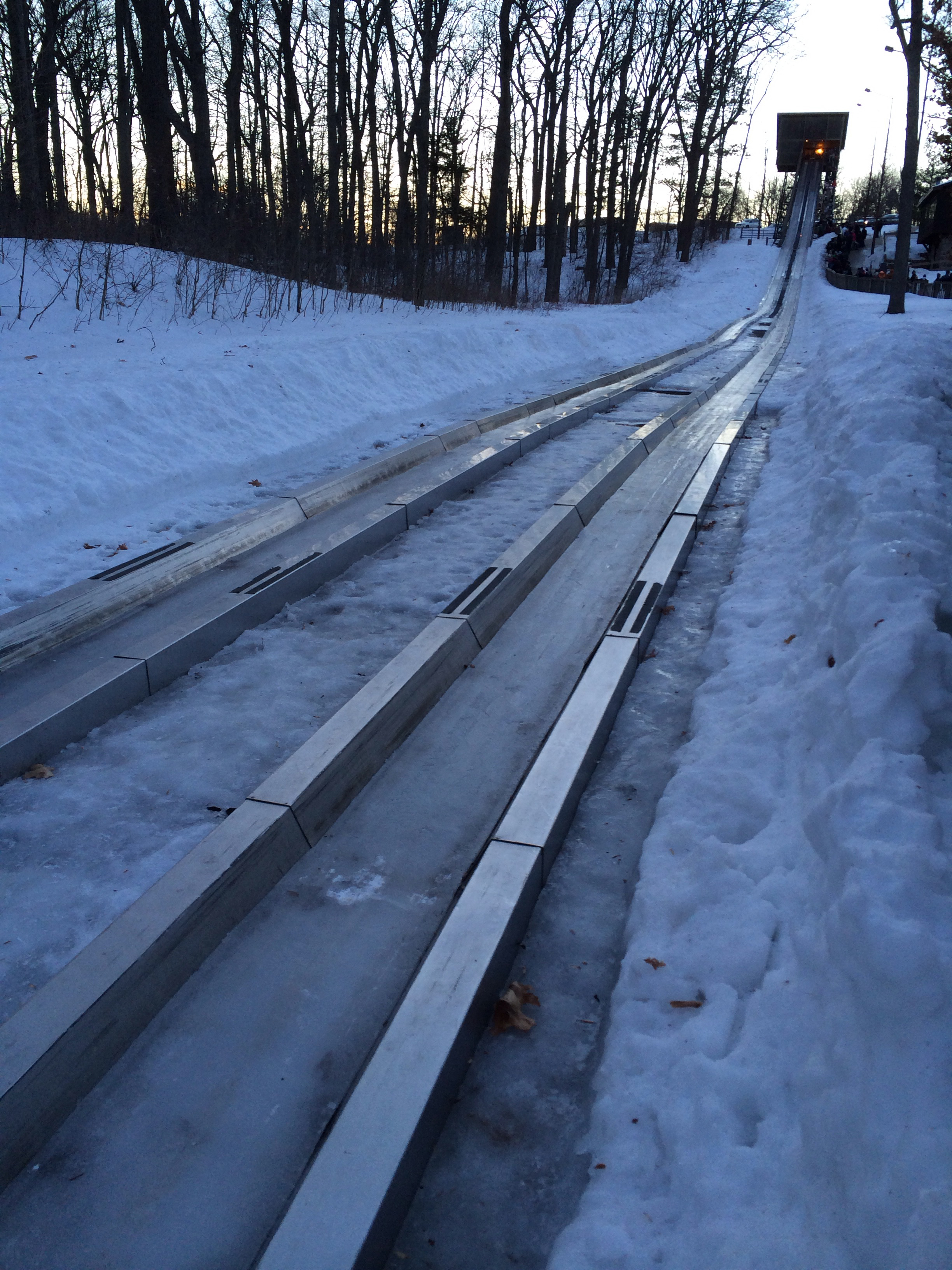 A snowy luge track with metal rails extends downhill, surrounded by trees, leading to a platform at the top.