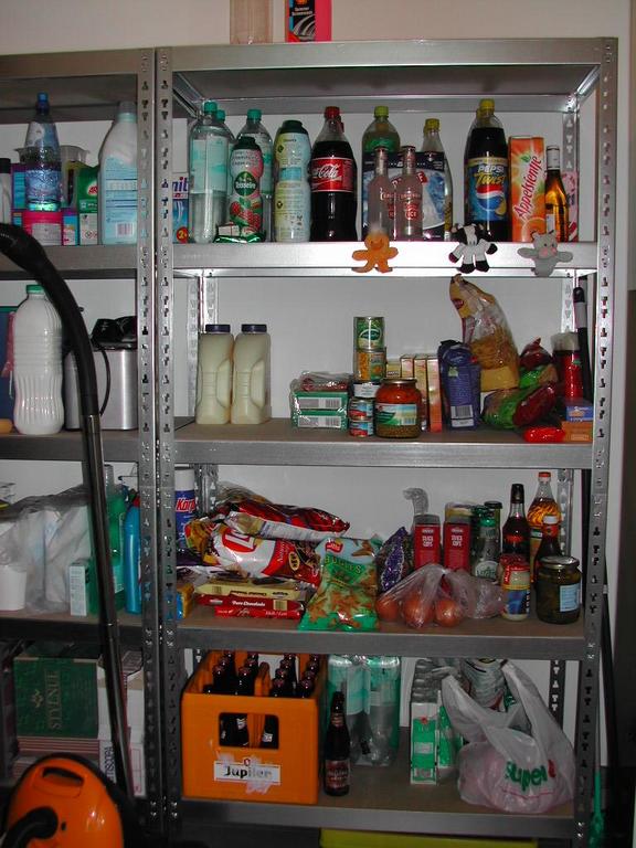 Metal shelves stocked with bottled drinks, food items, and household supplies in a kitchen, two months after moving in.