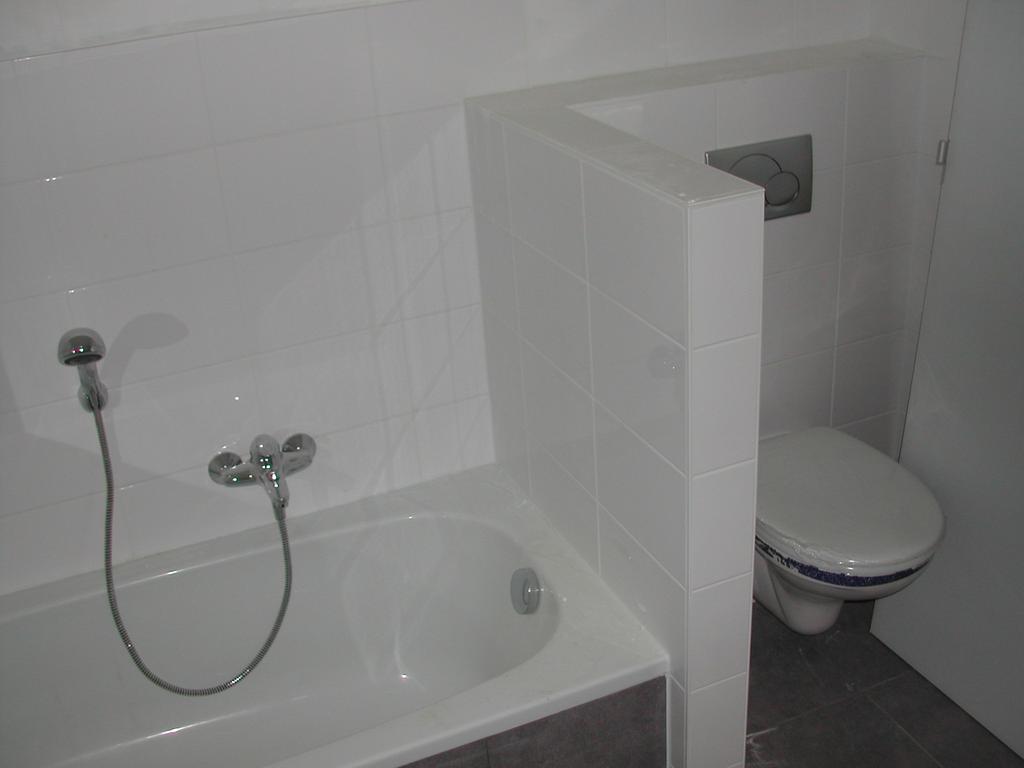 A bathroom with a bathtub, handheld shower, and a toilet partially hidden by a tiled partition wall.