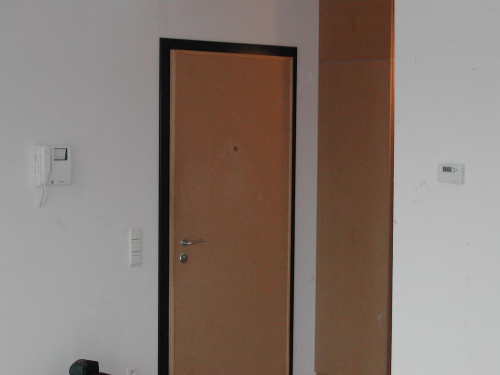 A plain living room with a closed wooden door, intercom, and thermostat on the wall.