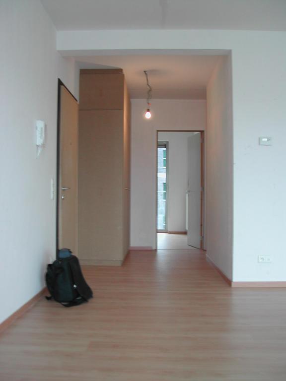 Empty living room with light wood flooring, white walls, and a black backpack on the floor near the door.