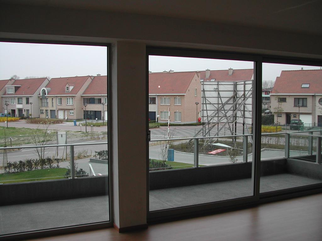 Empty living room with large windows and a balcony, showing a suburban street with houses and a billboard outside.